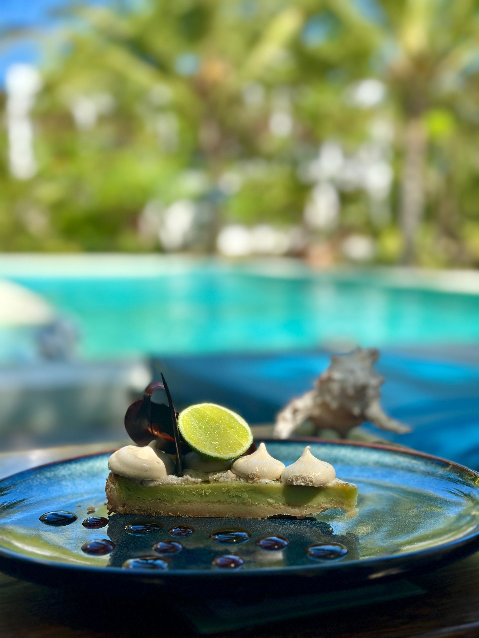 Lime tart dessert on blue plate by pool. Green filling, white cream, lime slice, tropical setting.