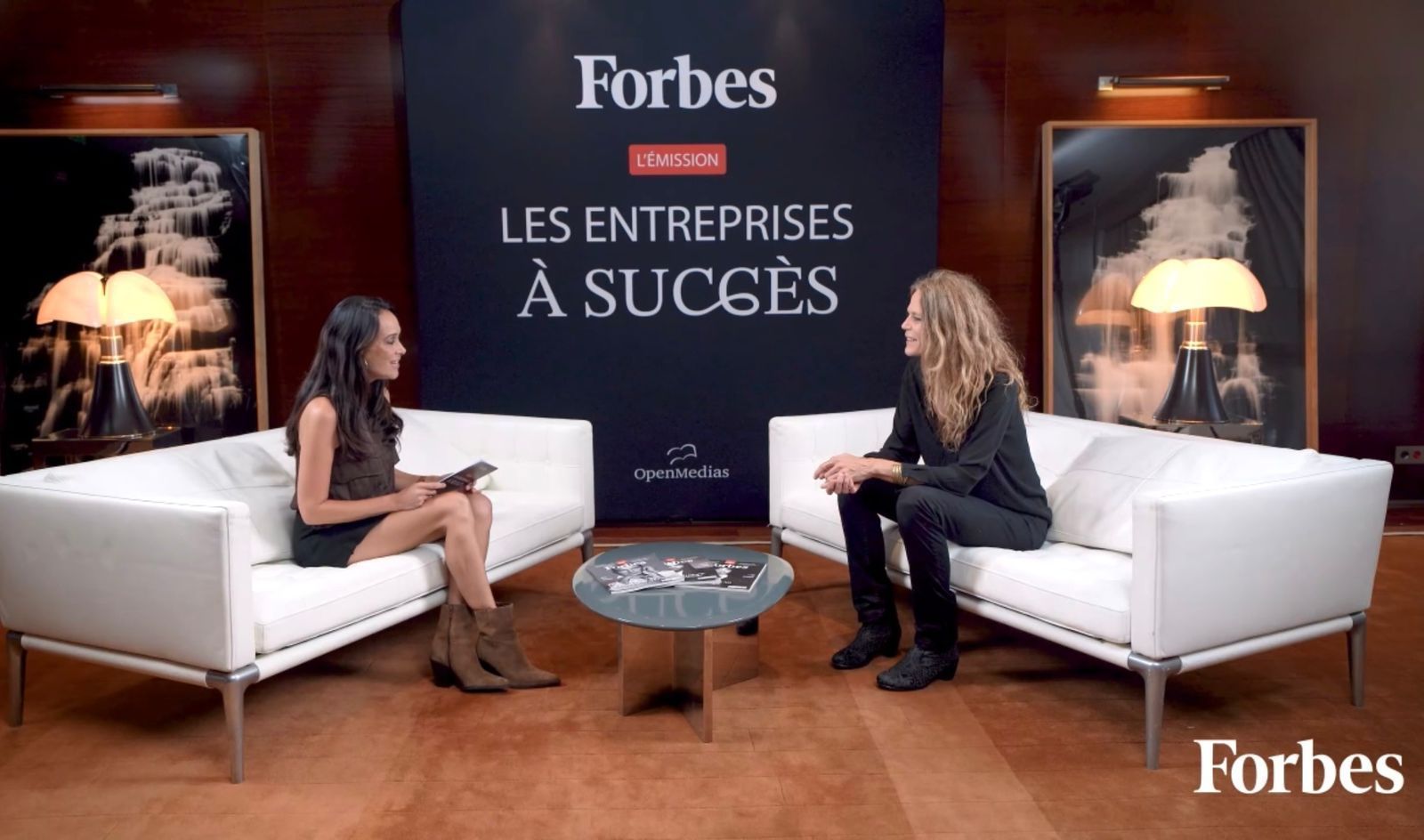 Two women seated on white couches, facing each other during a Forbes interview.