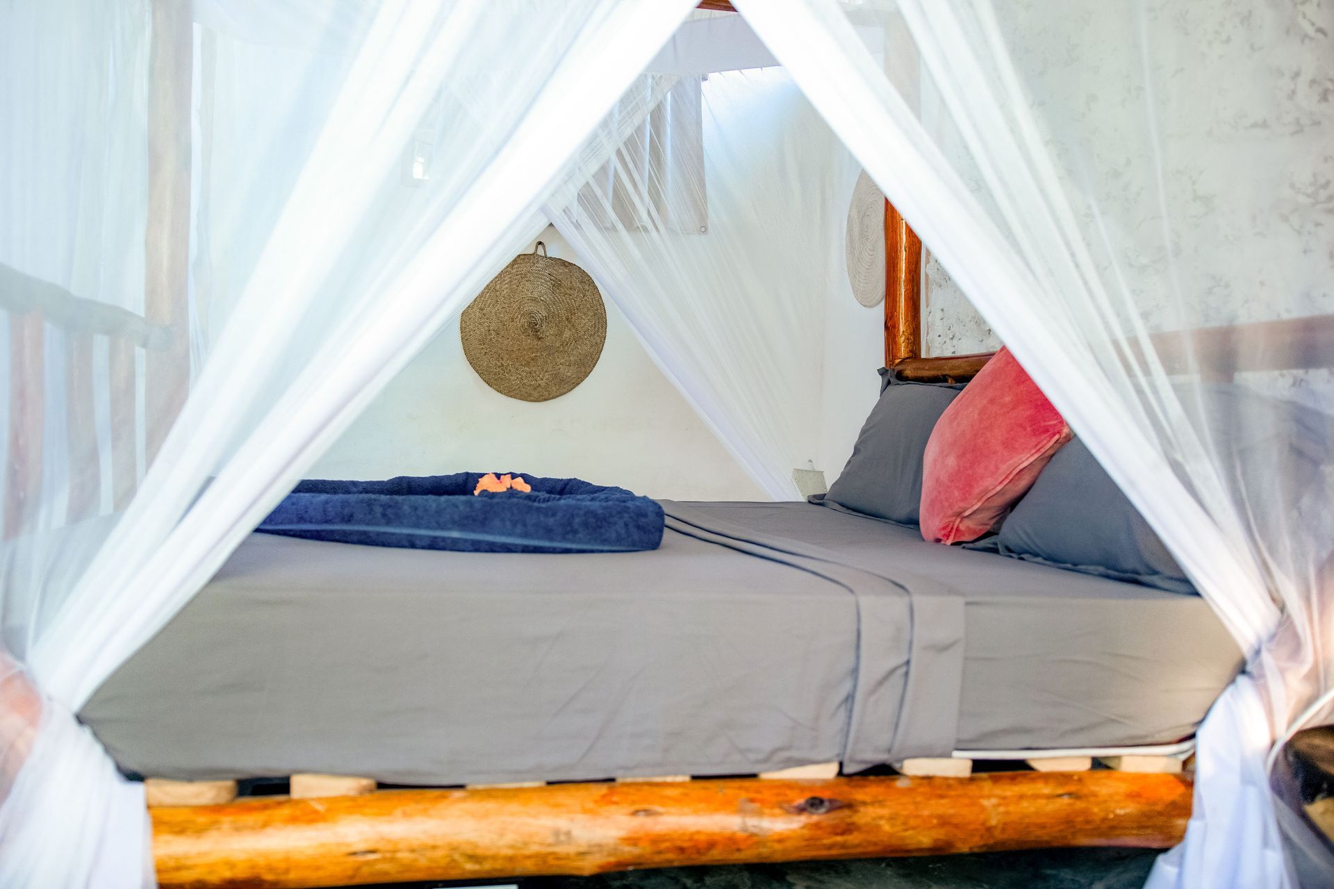 A canopy bed with grey sheets and a pink pillow; a blue towel sits on the bed.