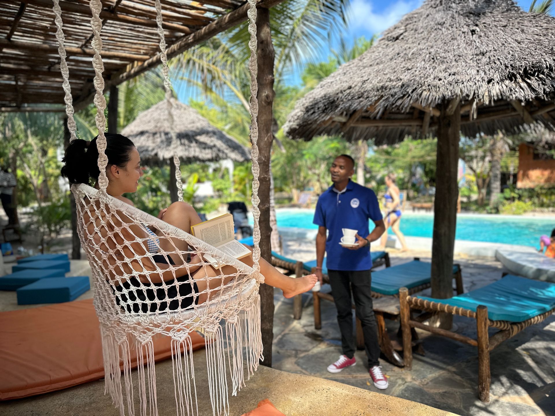 Woman in hammock reads book near pool; a staff member stands nearby, holding a cup.