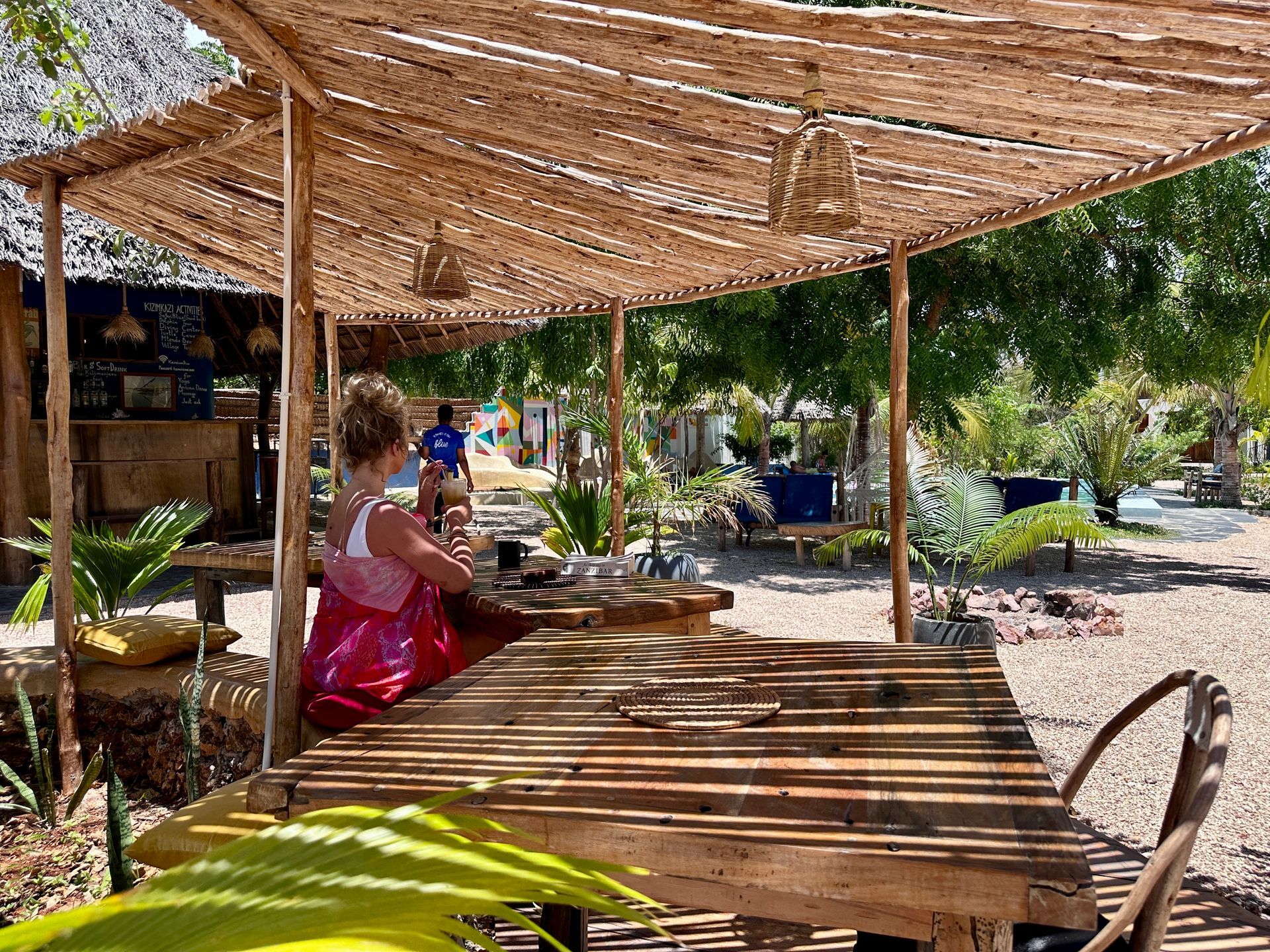 A woman is sitting at a wooden table under a thatched roof.