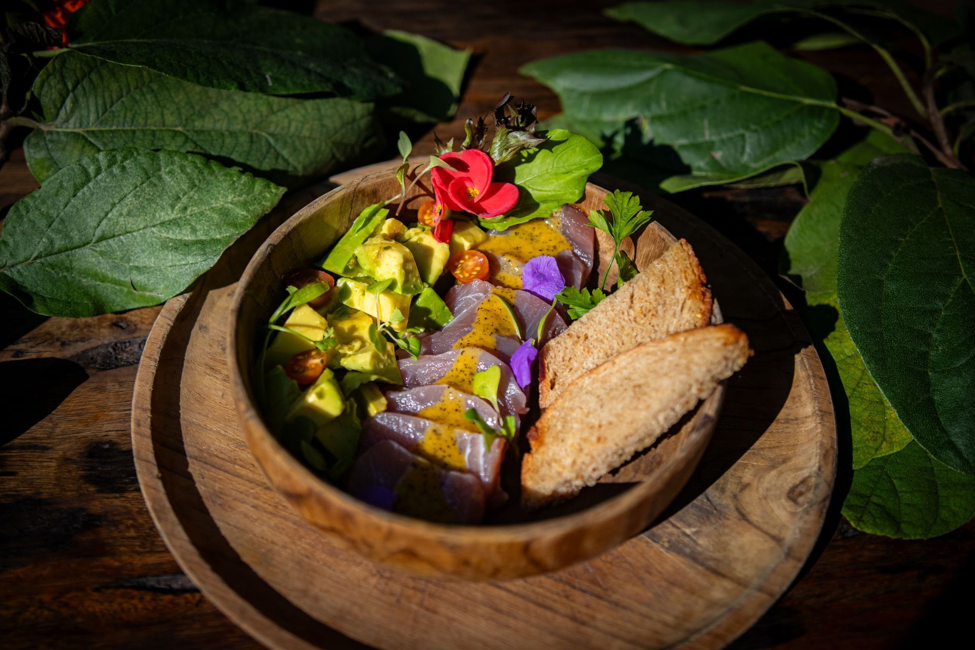 Wooden bowl of fresh tuna, avocado, and toast, garnished with flowers and leaves.