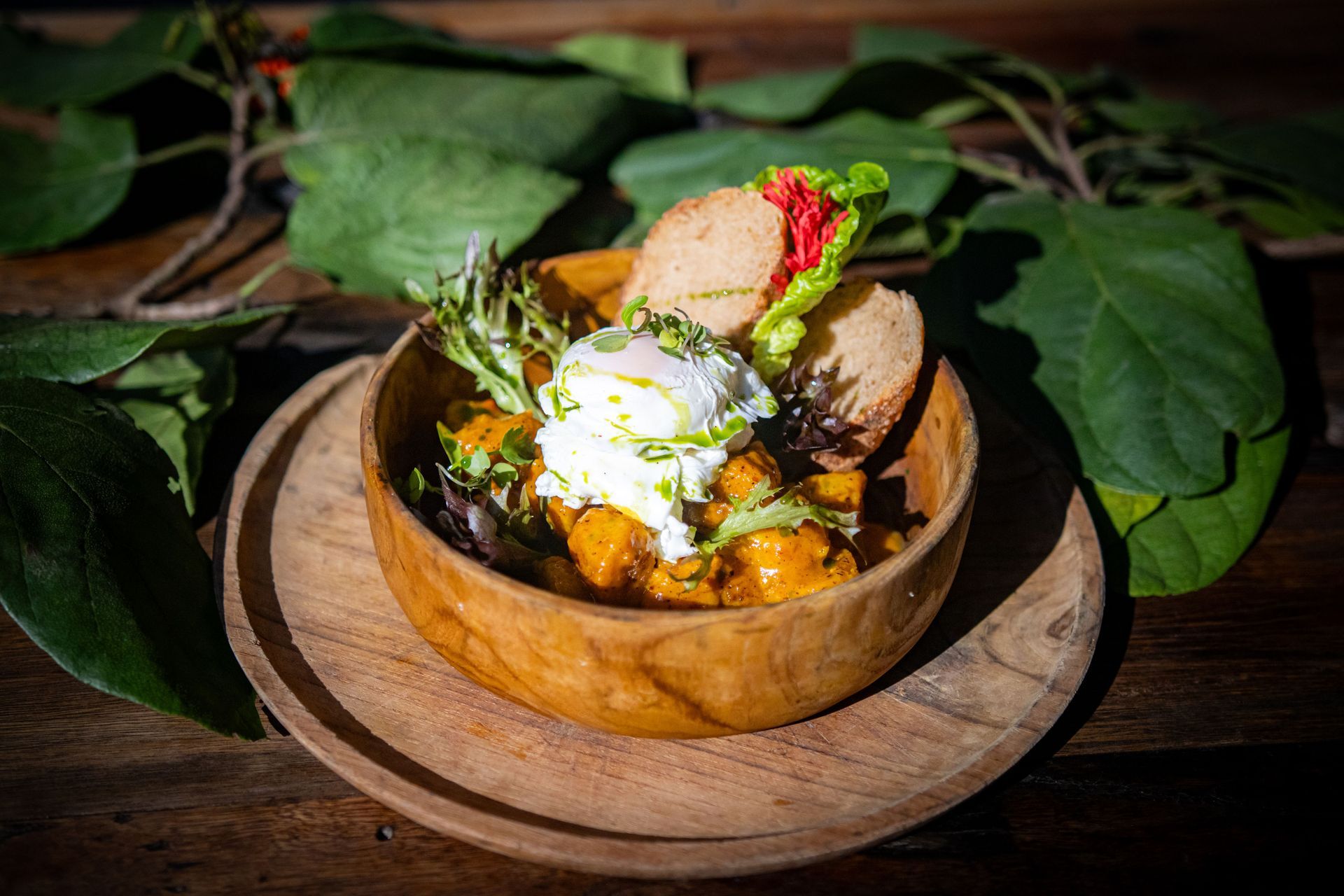 Bowl of food with poached egg, greens, and bread on a wooden plate, surrounded by leaves.