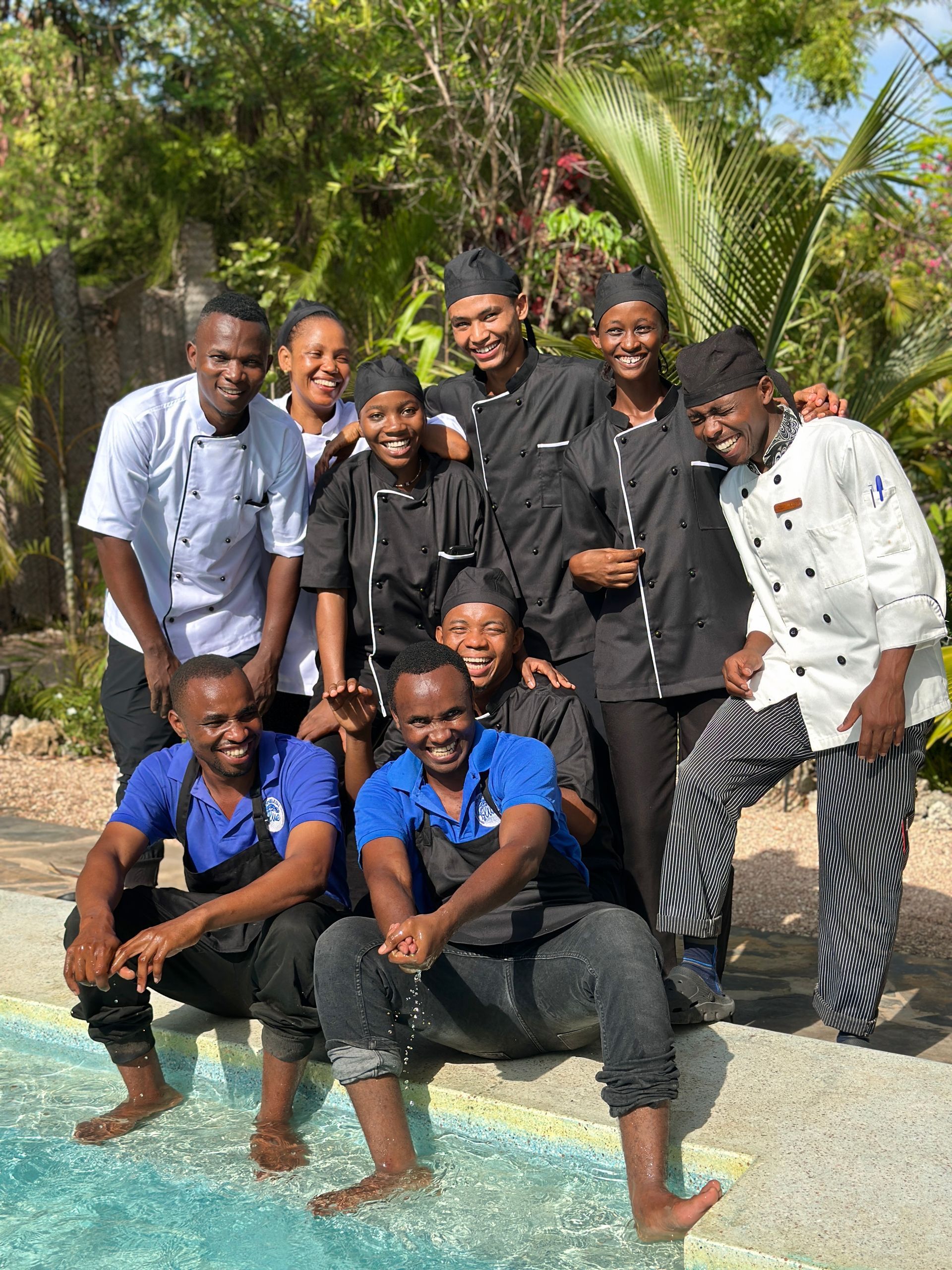 Group of culinary staff posing by a pool; some in chef whites and hats, others in blue aprons.