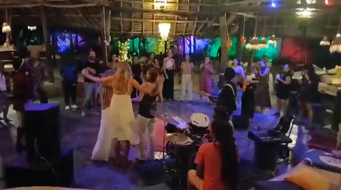 People dancing in a dimly lit indoor venue, with musicians playing nearby and colorful lights overhead.