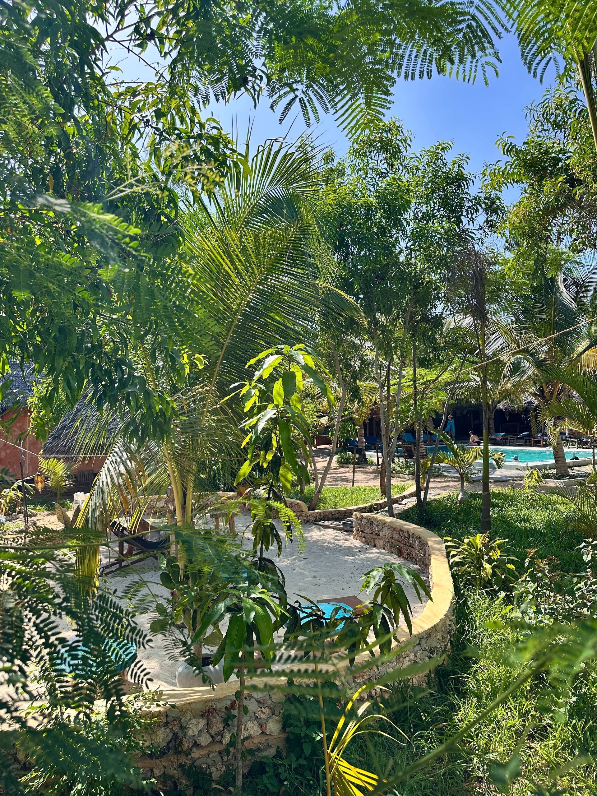Un espace extérieur luxuriant et verdoyant avec une structure circulaire en pierre, des arbres et une piscine au loin.