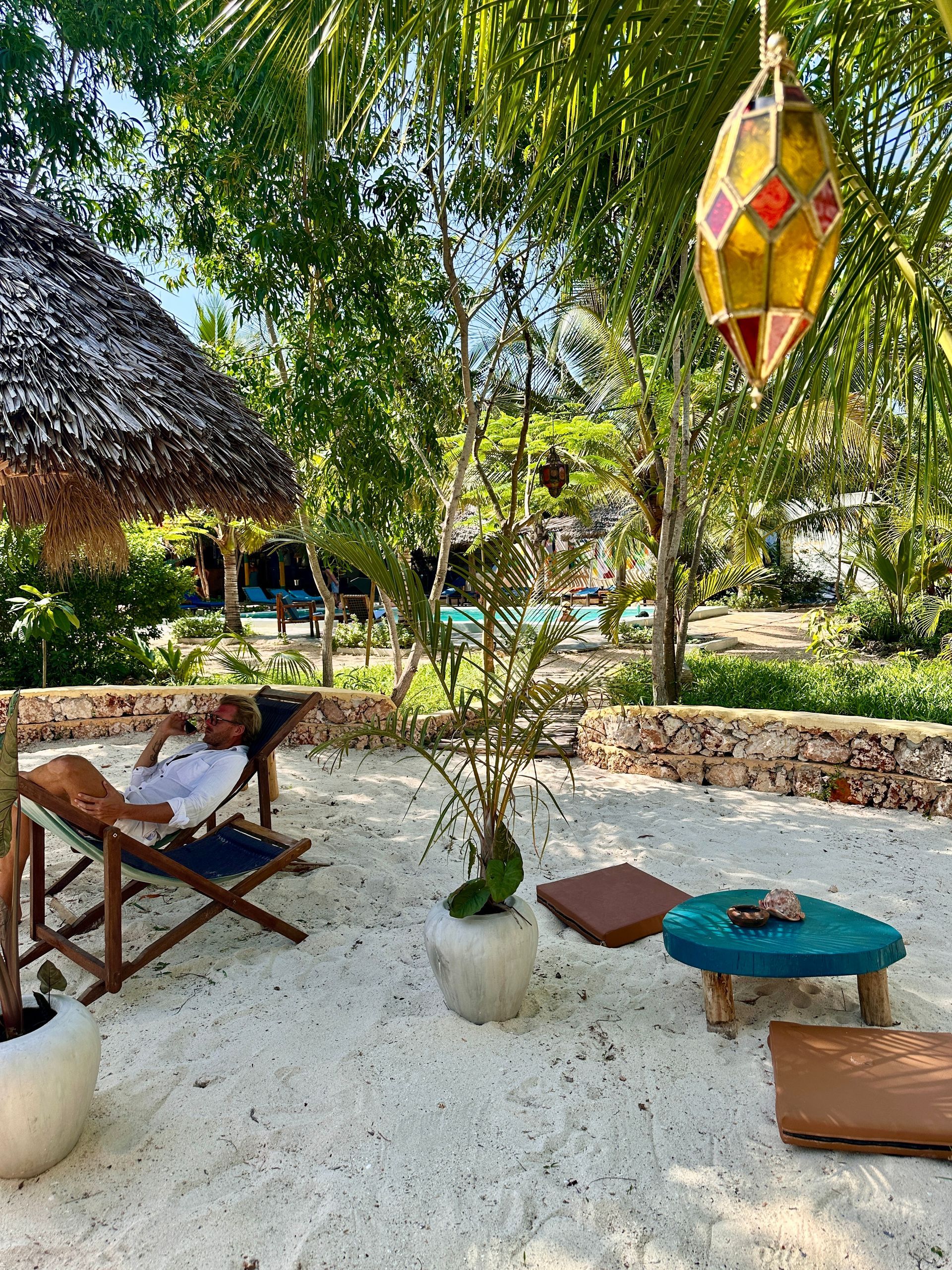 Un homme se détend sur une chaise de plage sous un toit de chaume, entouré de verdure et de coussins décoratifs.
