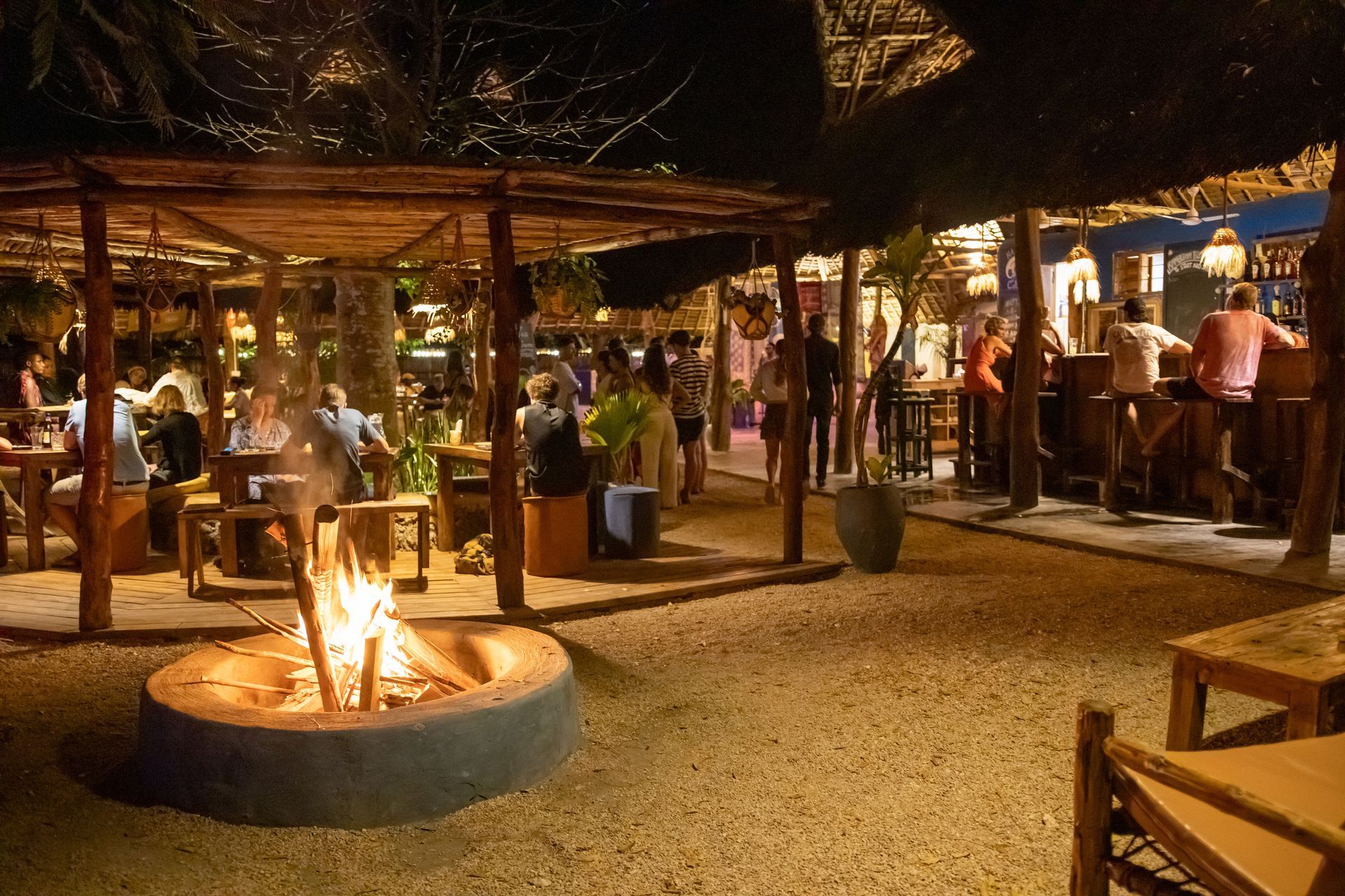 Outdoor bar at night with fire pit, people seated at tables and at the bar, under a thatched roof structure.