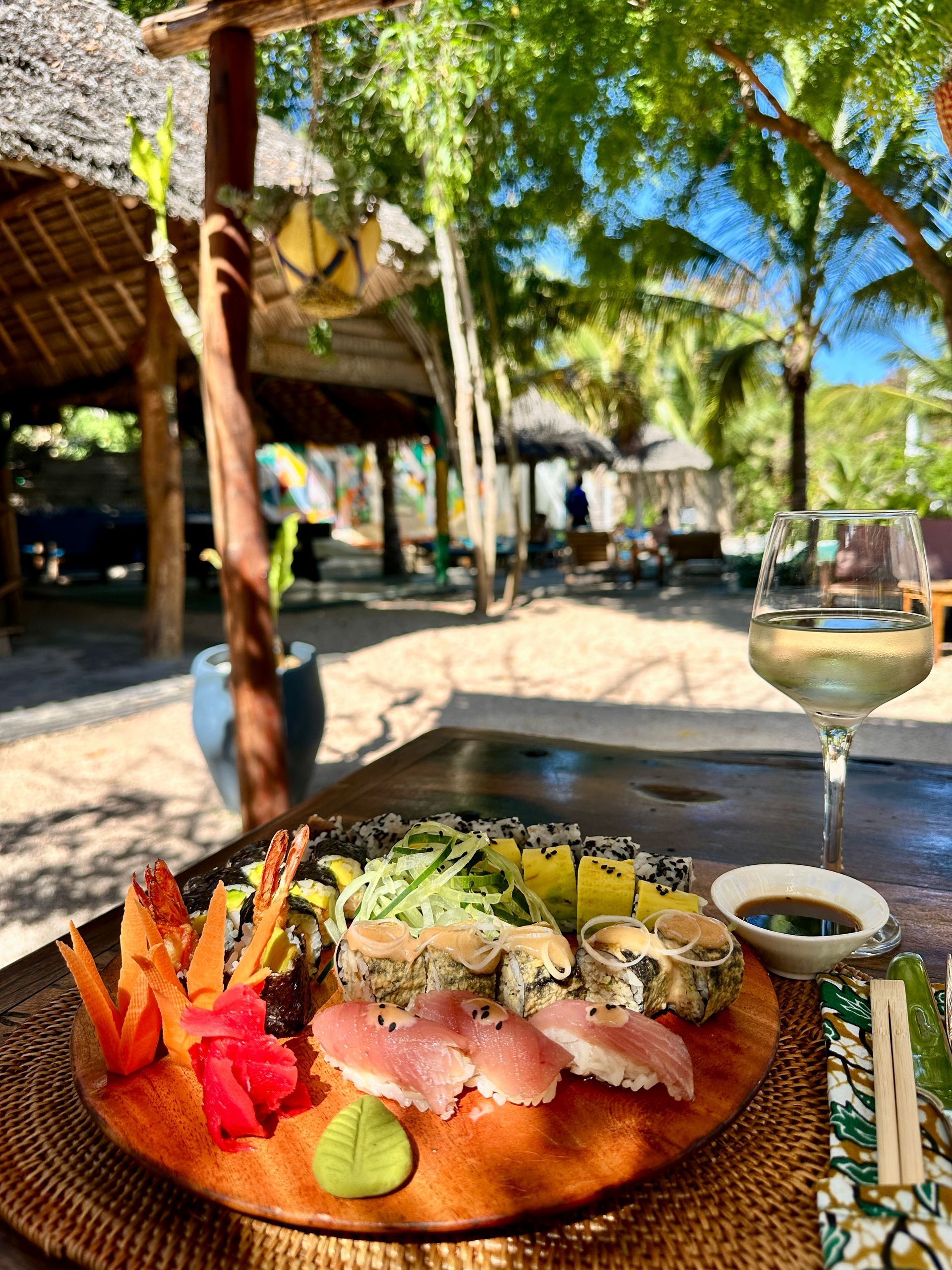 Sushi and wine on a wooden platter at an outdoor restaurant with thatched roof structures and palm trees.