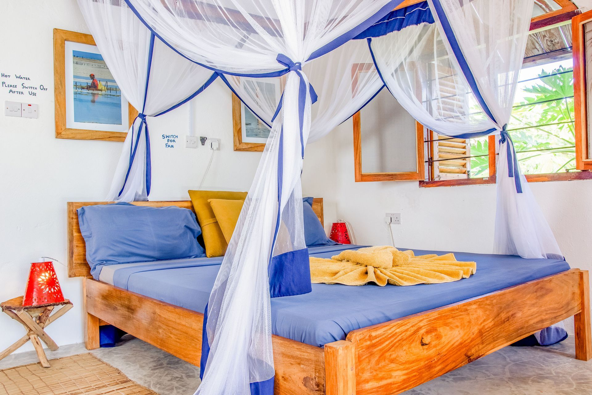 Bedroom with wooden bed, blue linens, mosquito net, artwork, and small table.