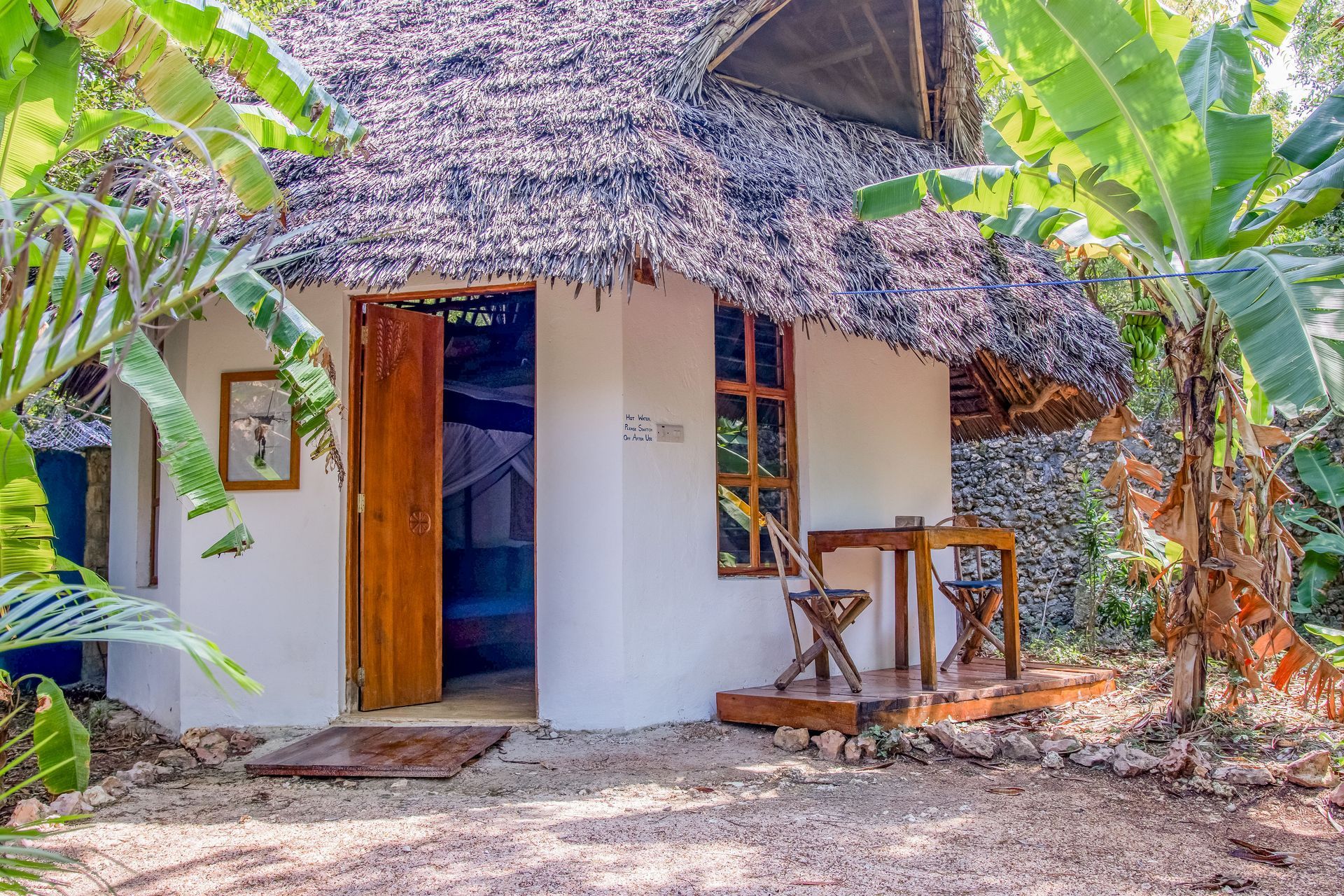 Small white tropical cottage with thatched roof, wooden door and window, surrounded by greenery.