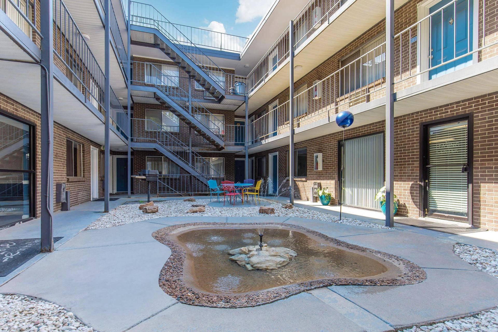 Courtyard with three-story apartment building and a small pond with fountain. Metal staircase and outdoor seating present.
