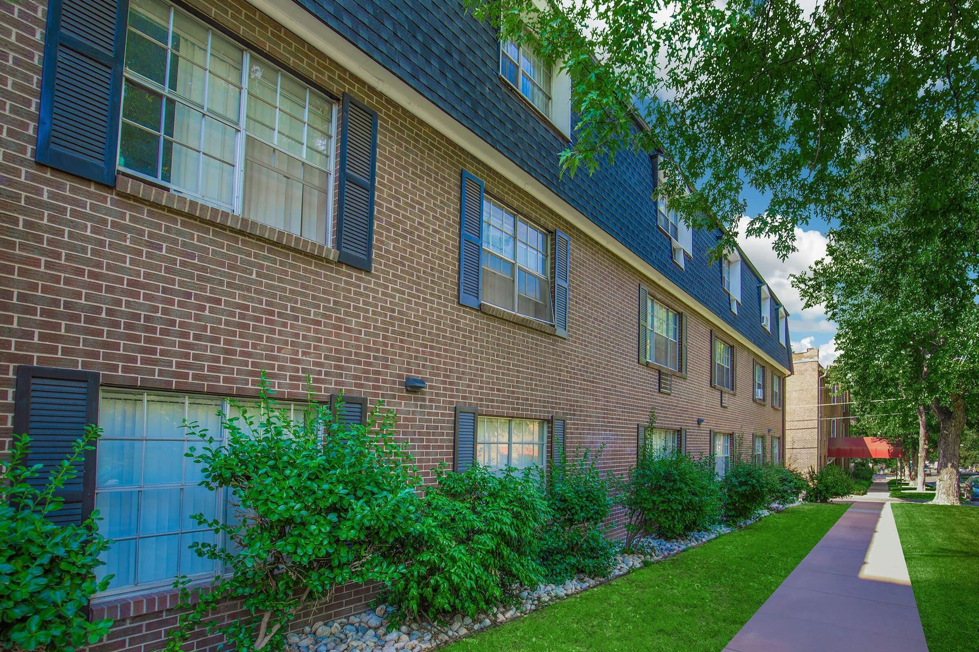 Brick apartment building with dark shutters and roof, green bushes, sidewalk, and trees.