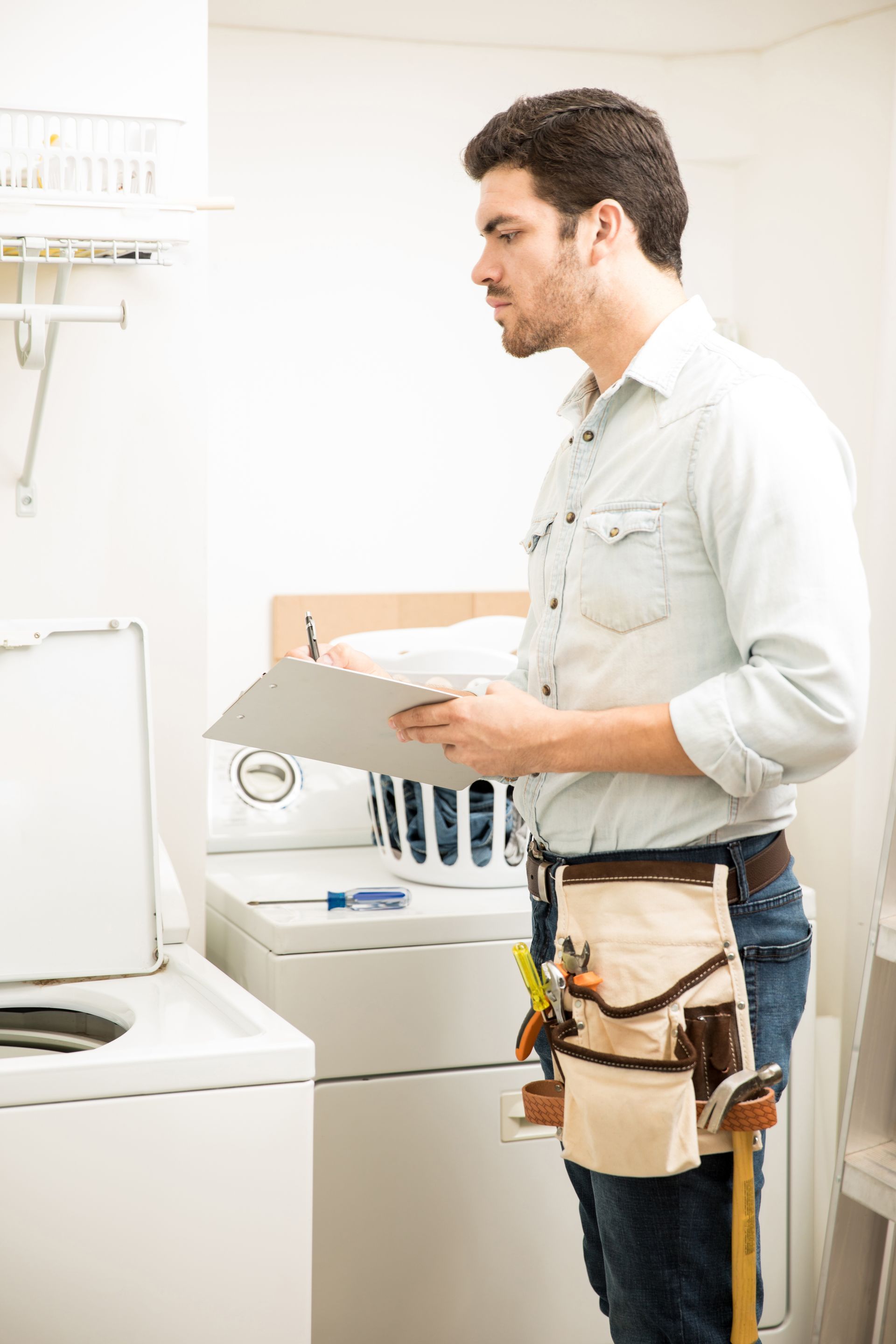 Technician holding a clipboard in a laundry room beside a washing machine and utility sink
