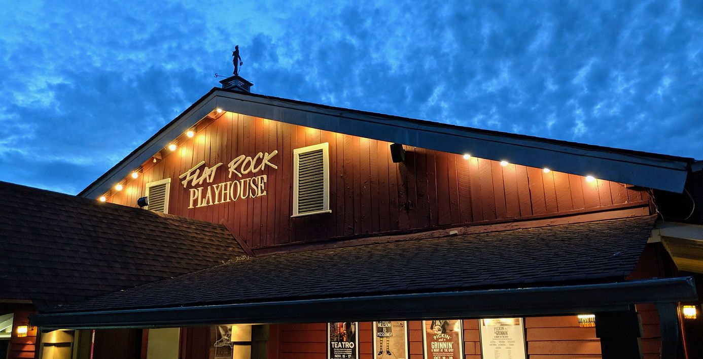 Exterior of the Flat Rock Playhouse at dusk, with illuminated sign and string lights against a dark wood building.