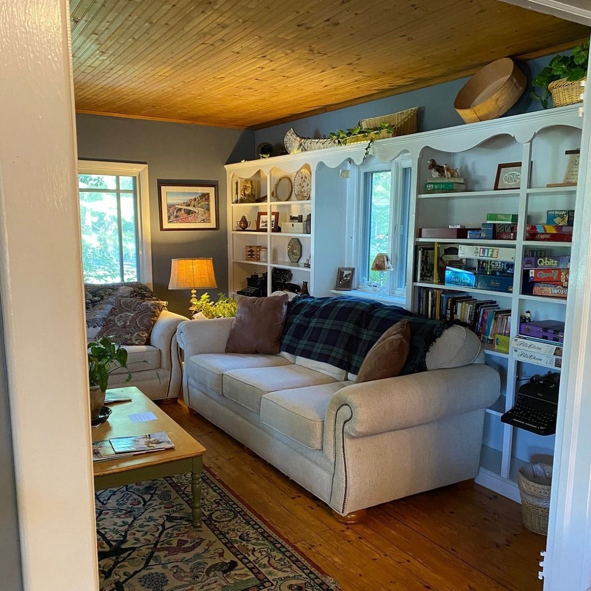 Cozy living room with white bookshelves, a beige sofa, and wooden floors. Blue walls and a wooden ceiling add to the warm atmosphere.