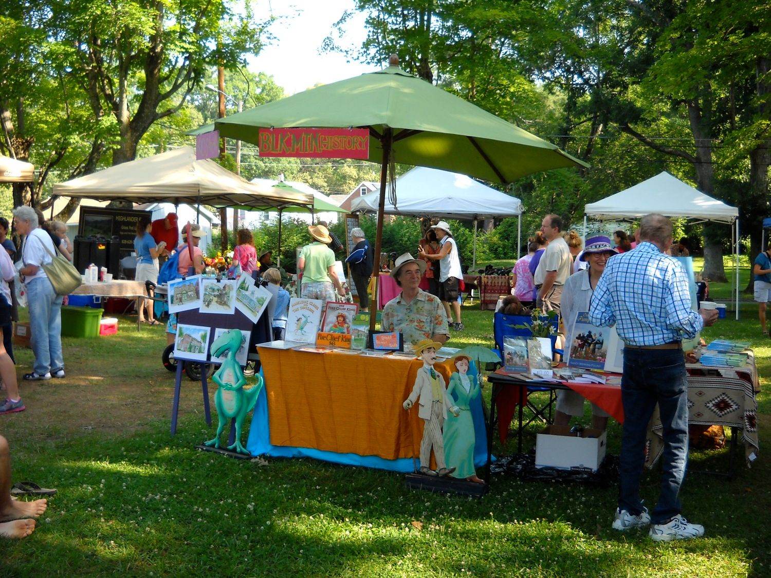Outdoor art fair with vendors under tents in a grassy park. People browse artwork and interact with vendors on a sunny day.