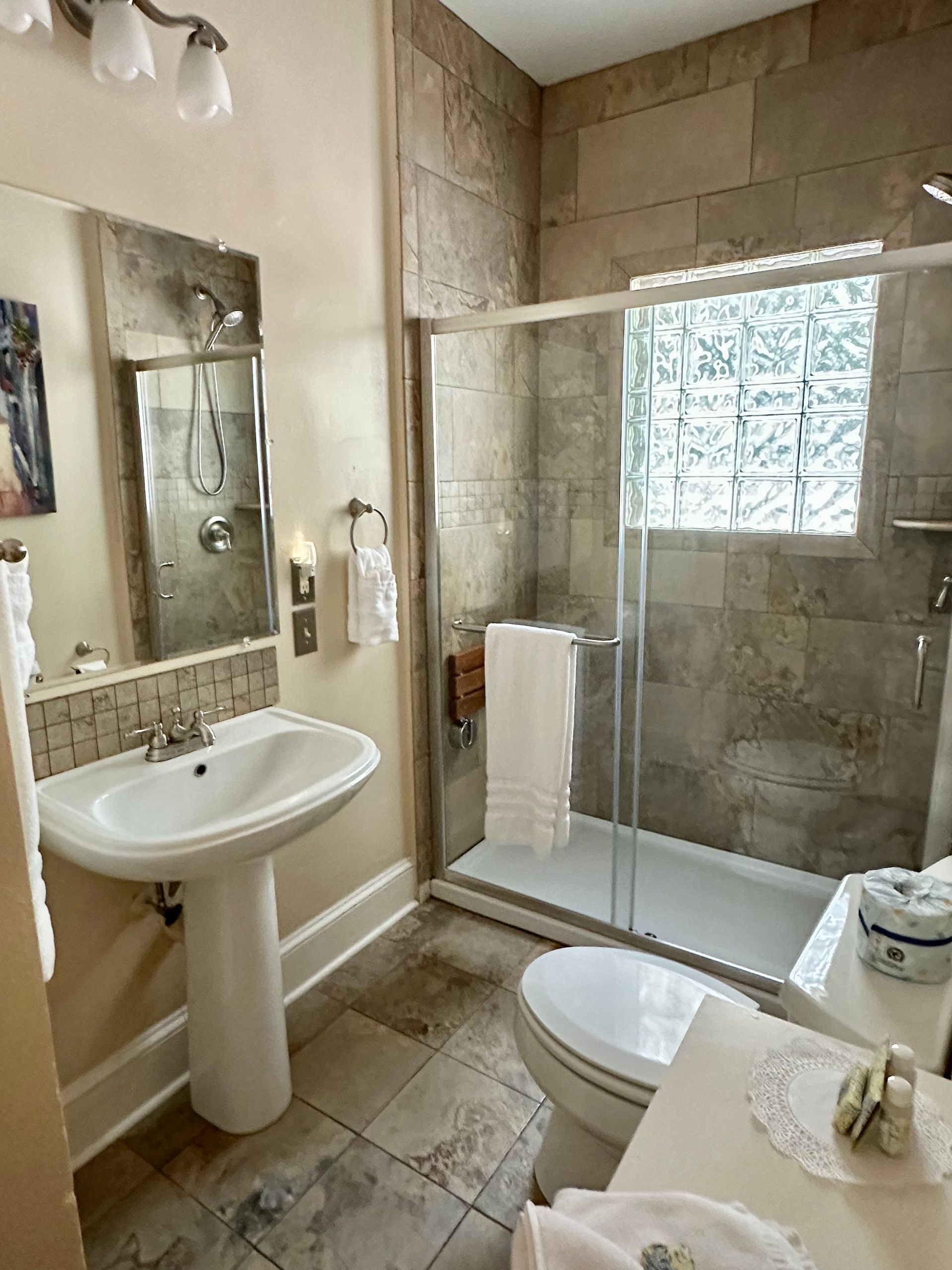 Bathroom with pedestal sink, tiled shower, and glass block window; neutral tones, light fixtures.