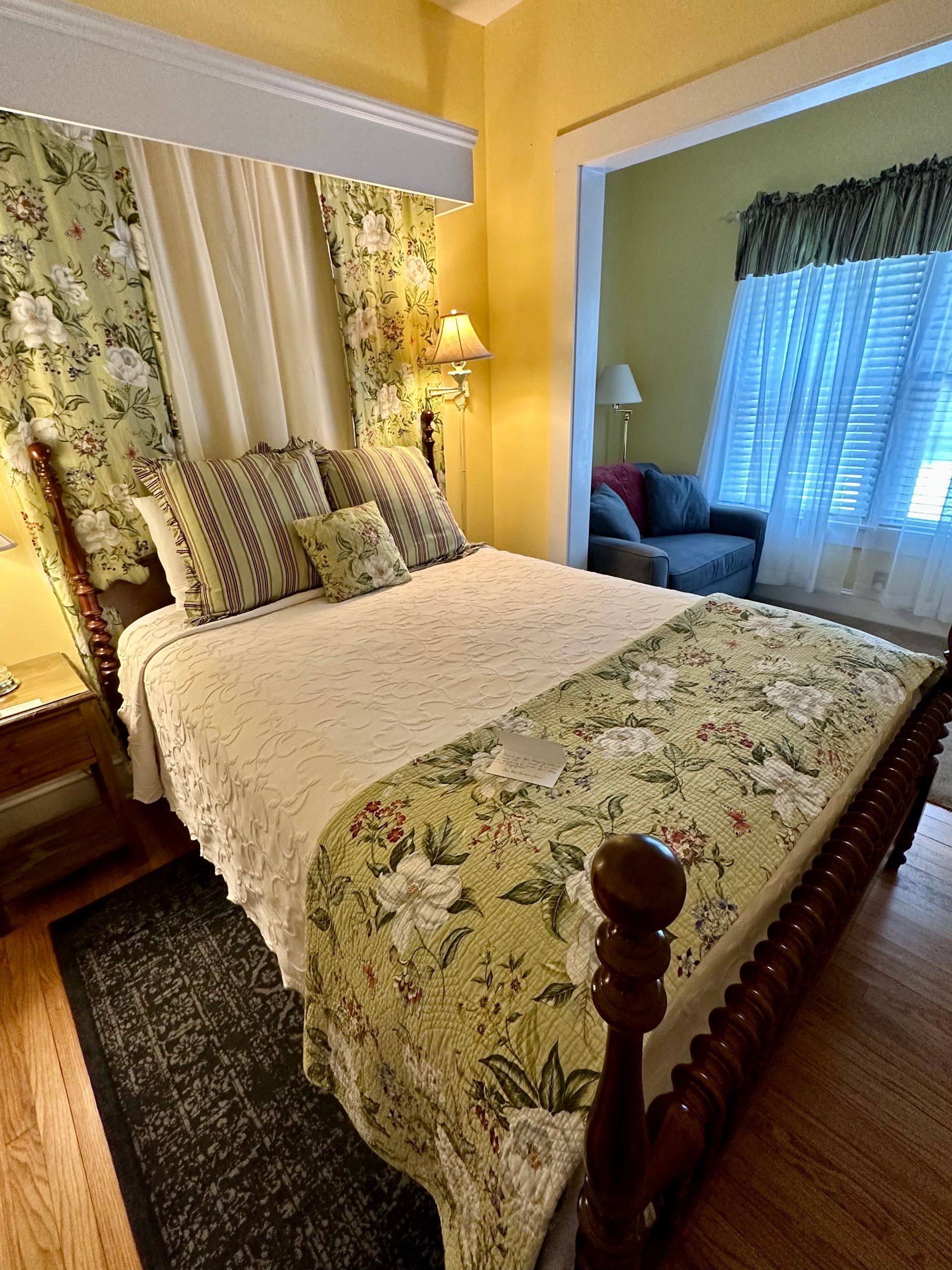 A bedroom with a four-poster bed covered in floral bedding. An open doorway reveals a sitting area with a couch.