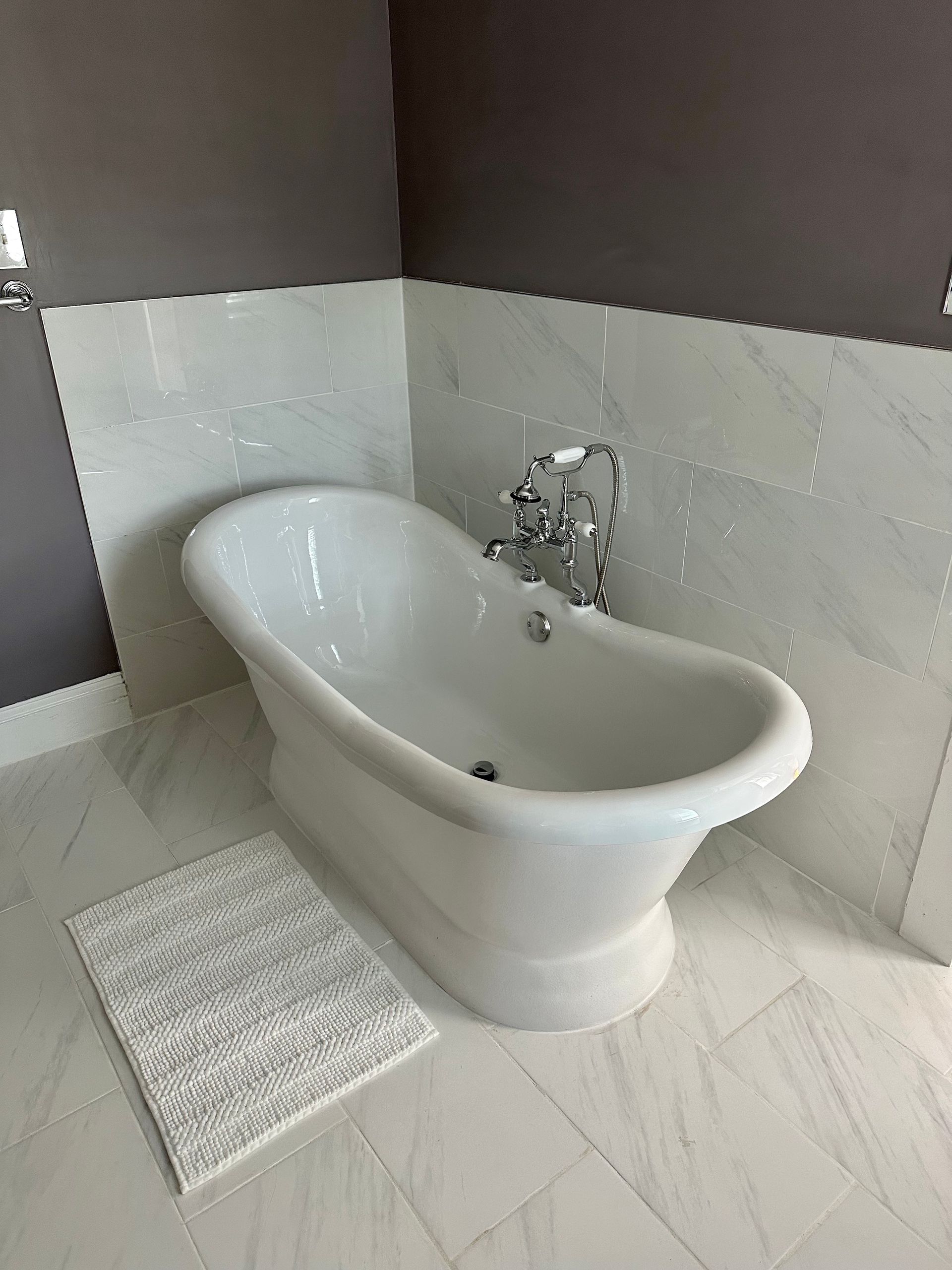 A white, classic claw-foot bathtub sits against marble tiled walls. A bath mat lays beside the tub.