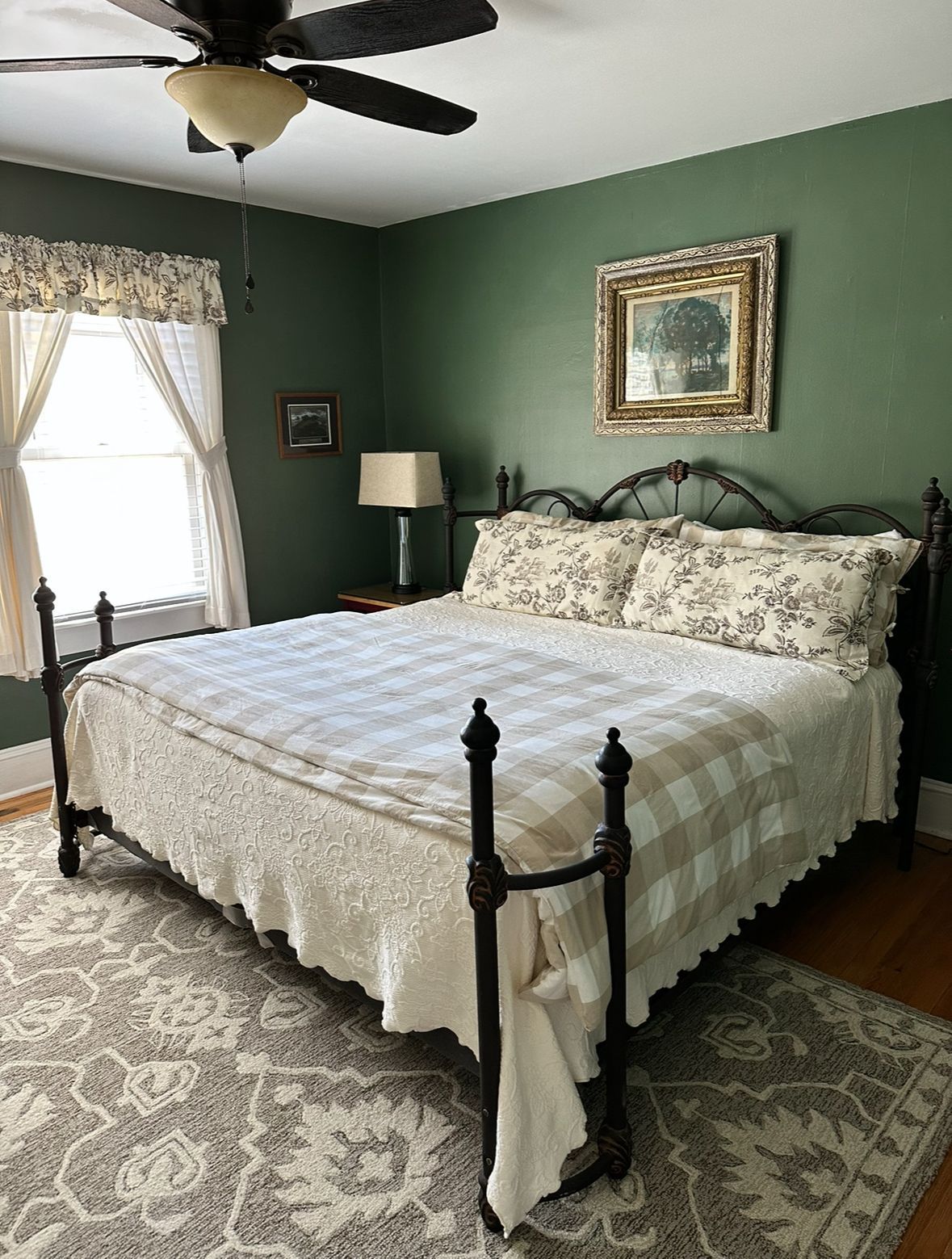 A bedroom with a wrought-iron bed, green walls, and a floral-patterned quilt. A framed painting hangs above the bed.