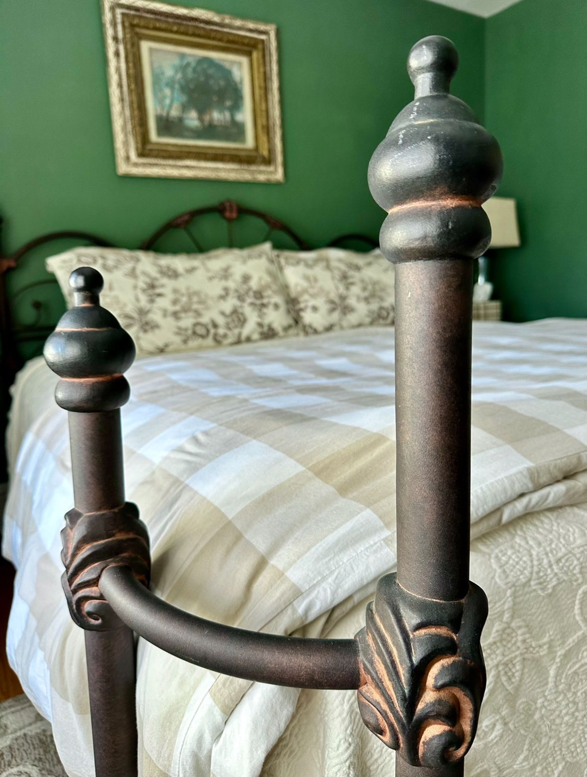 Close-up of a decorative metal bed frame with a green wall and framed art in the background. The bed is made with a light-colored plaid comforter.