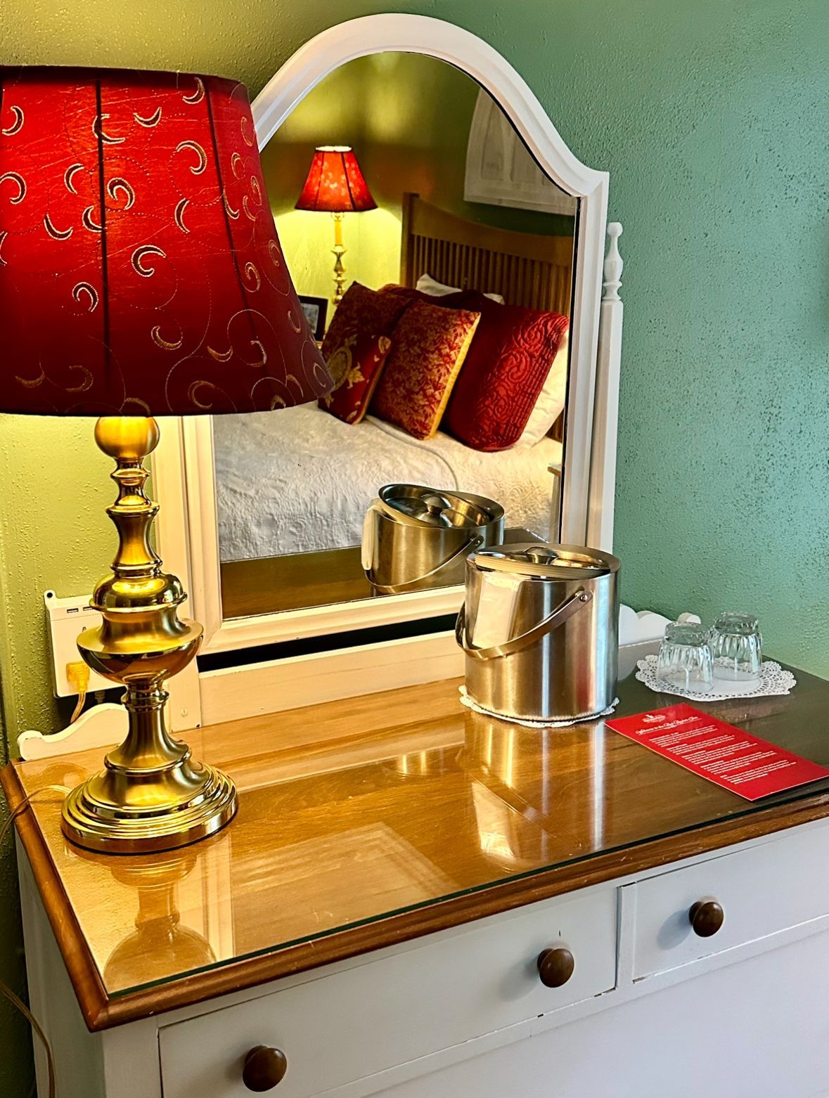Dresser with a red lampshade, a mirror reflecting a bed with pillows, and an ice bucket. Green and white walls.