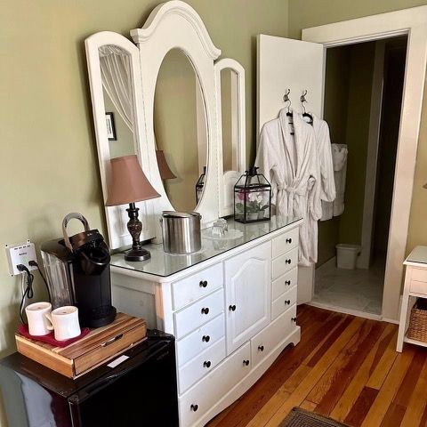 A white dresser with a large mirror, coffee maker, and open door to a bathroom with a bathrobe.
