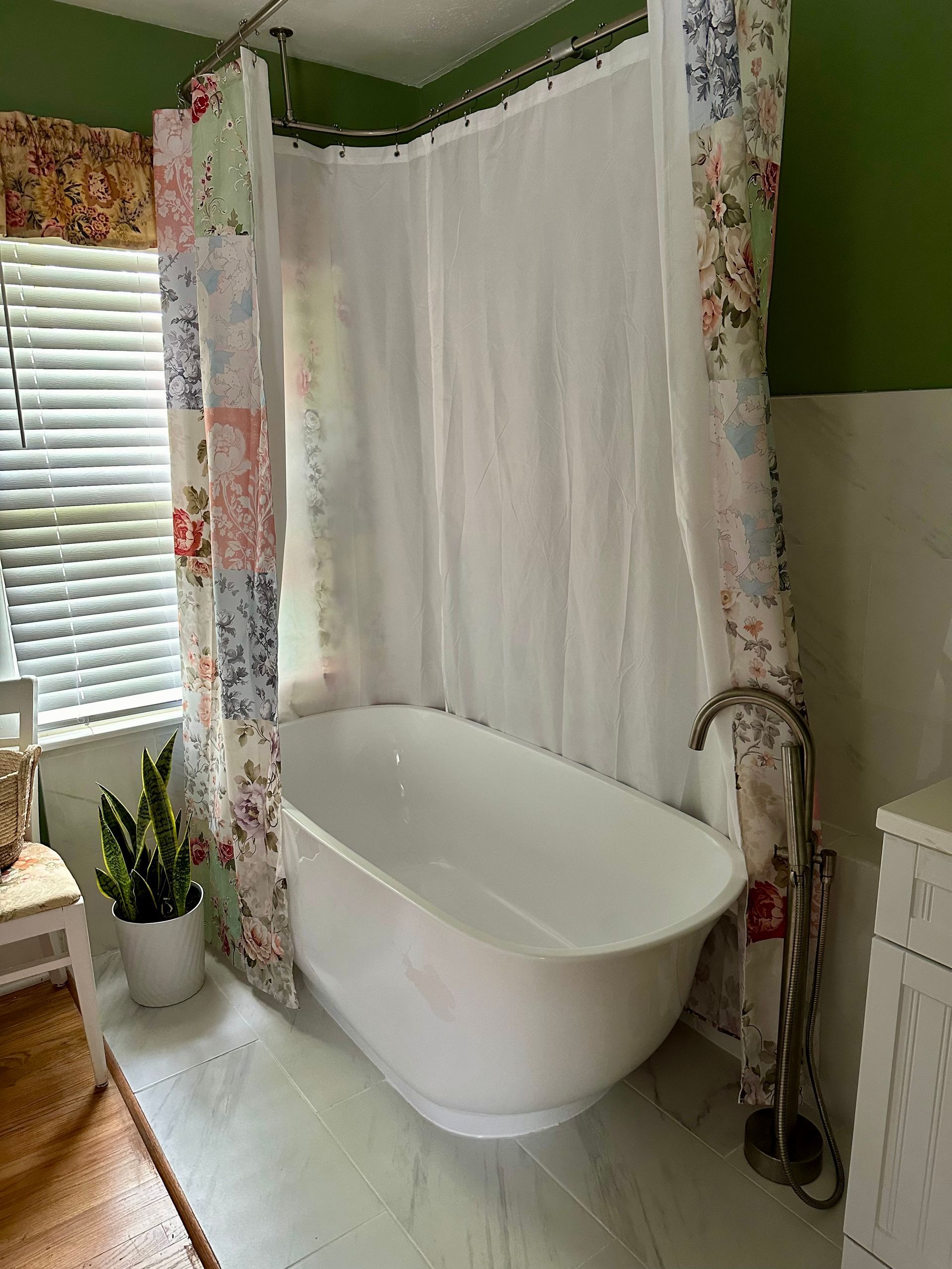 A white freestanding bathtub inside a bathroom, with a floral shower curtain and window blinds. Green and white walls.