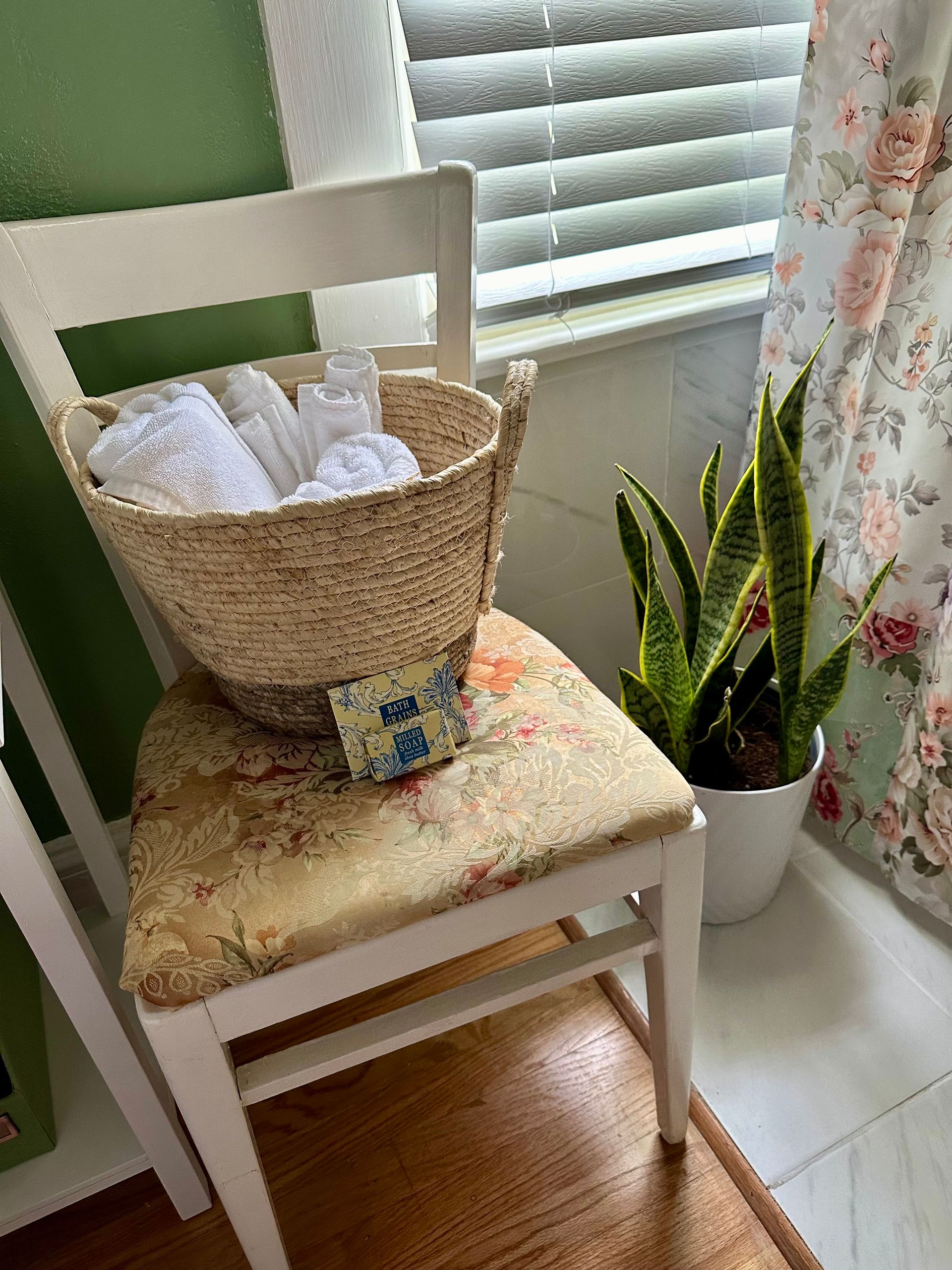 White chair with basket of towels and toiletries, snake plant in white pot, and floral curtain.
