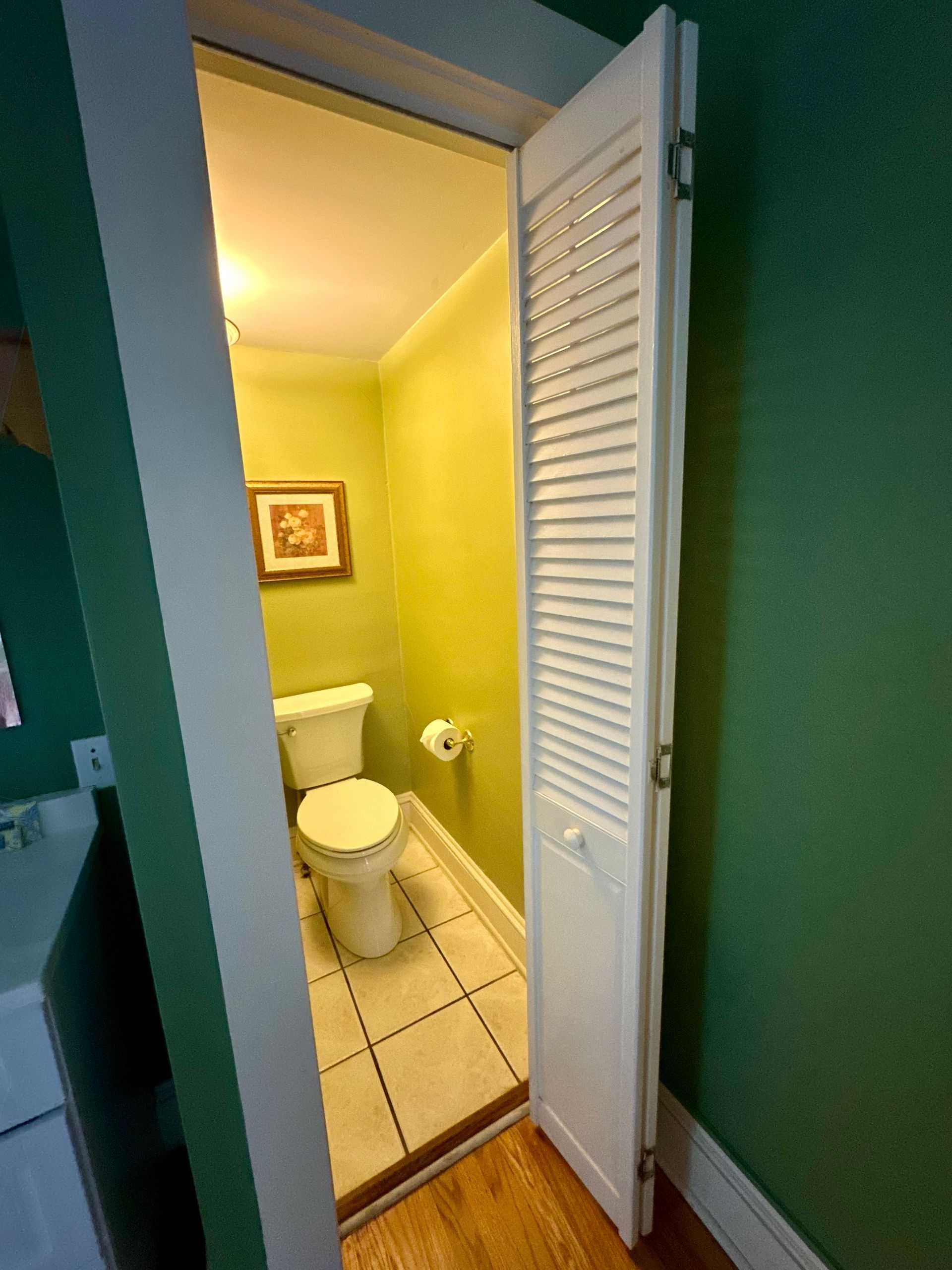 A small, brightly lit half bathroom with white louvered doors. The room features a toilet, artwork, and green walls.