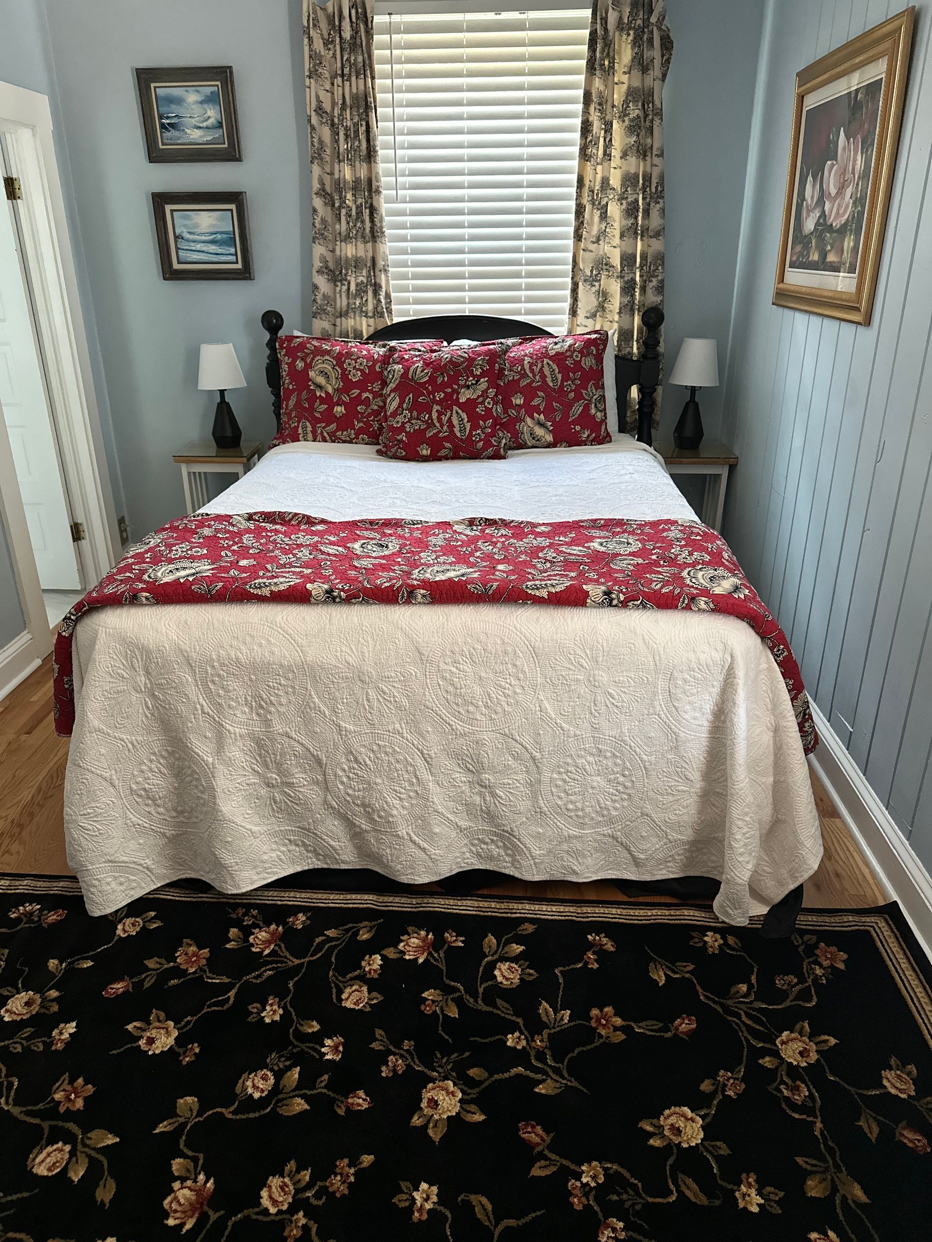 Small bedroom with blue walls, wooden floor, and a bed with red floral bedding. A window with patterned curtains is above the bed.