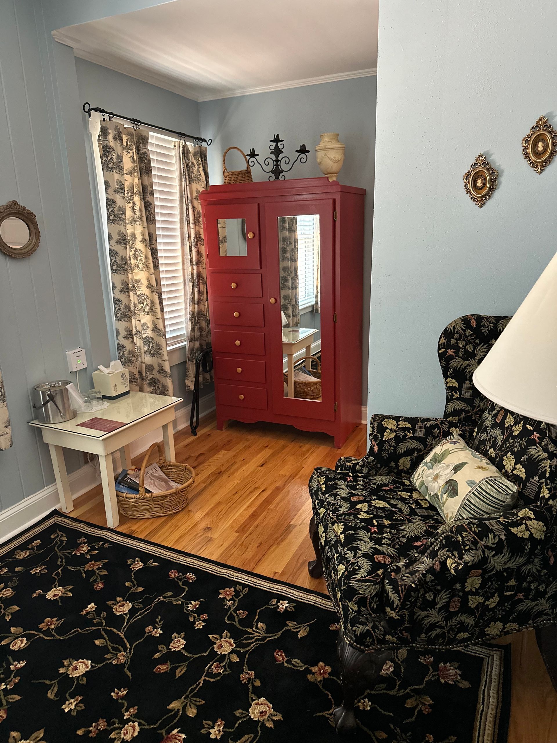A room with a red wardrobe, floral chair, and desk with a black floral rug. The walls are light blue.