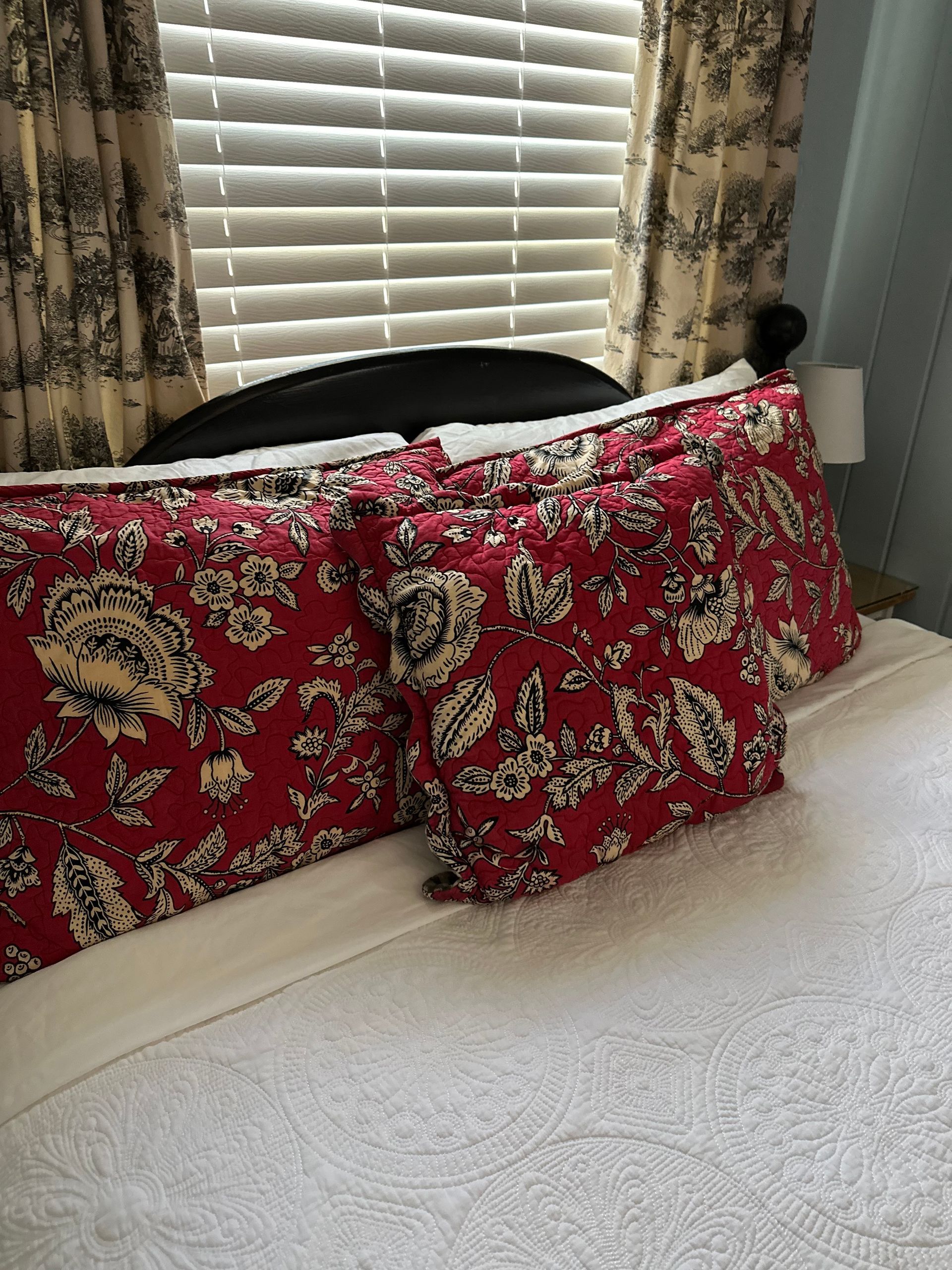 Bed with red floral pillows and curtains; window with blinds in the background.