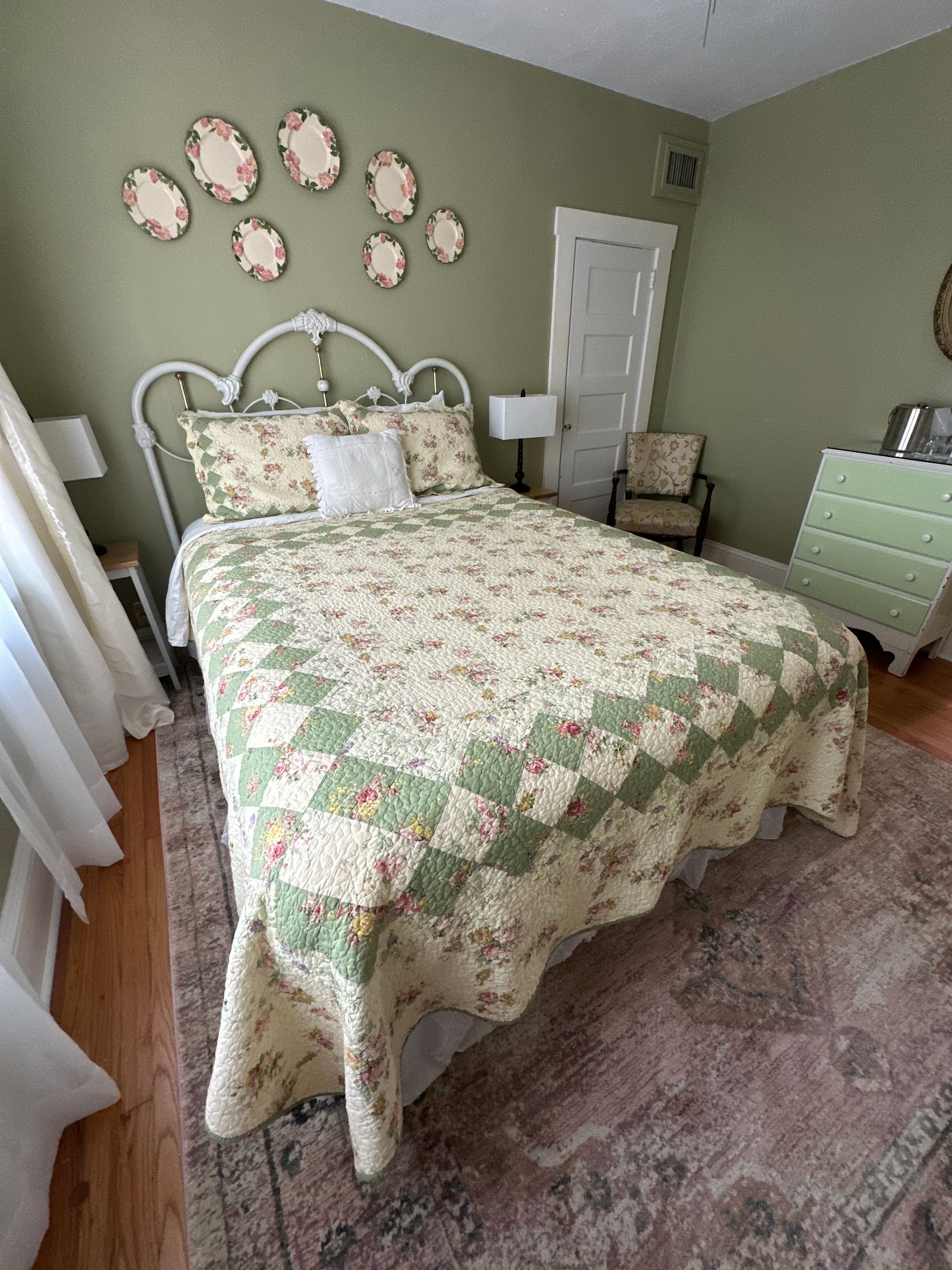 Cozy bedroom with white bed, floral bedding, chair, and dresser. Light green walls, and decorative plates.