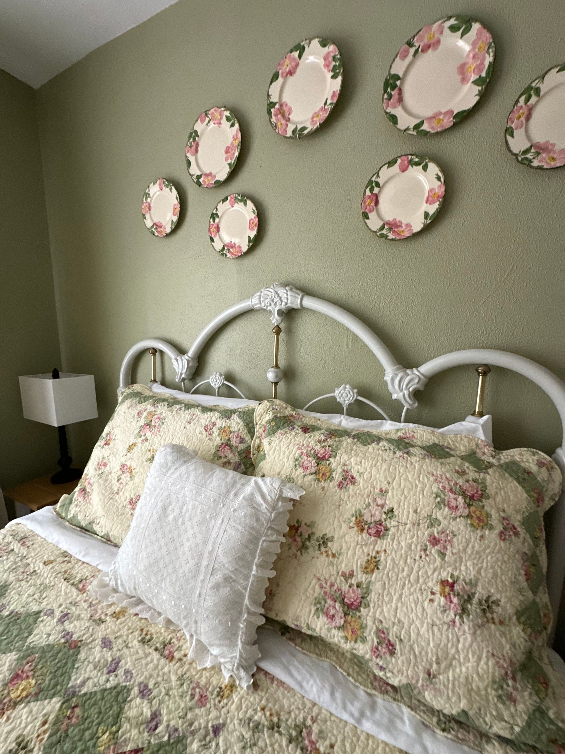 Bedroom with light green walls. A tan wingback chair holds a red throw blanket. A lamp sits on a side table next to a bed, and another stands behind the chair.