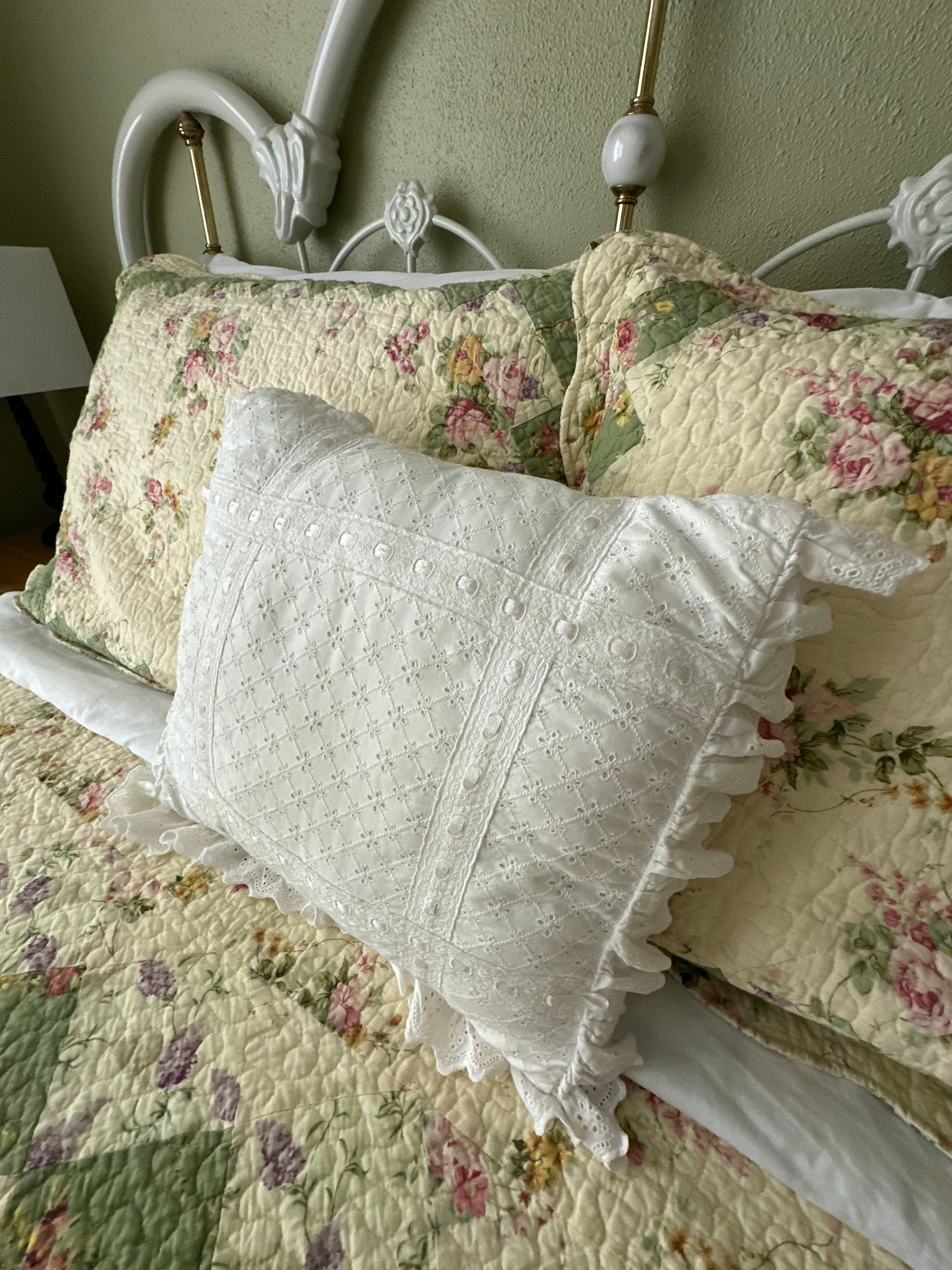 Bedroom with a white wrought-iron bed, floral bedding, a lamp, and a comfy chair. The walls are light green, and the room is warmly lit.