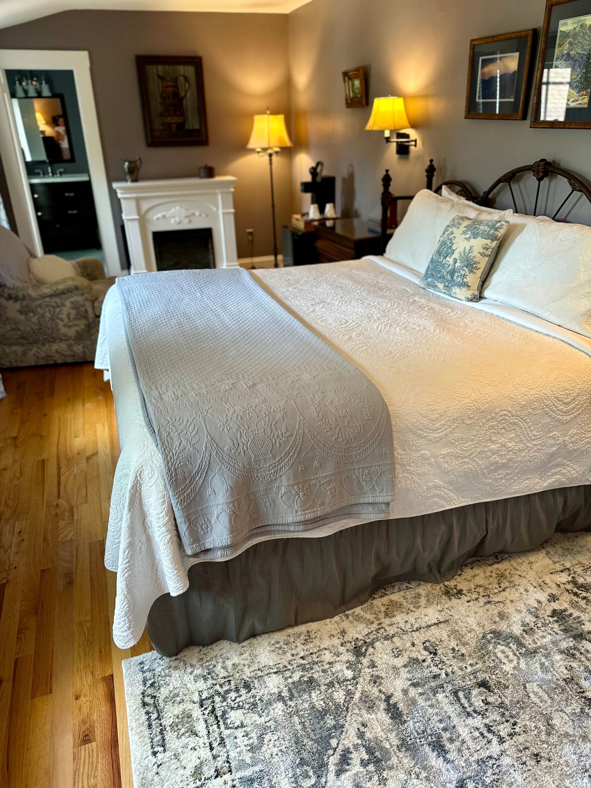 A bedroom with a bed, a fireplace, and a side table. Hardwood floors are visible, and the walls are painted gray.