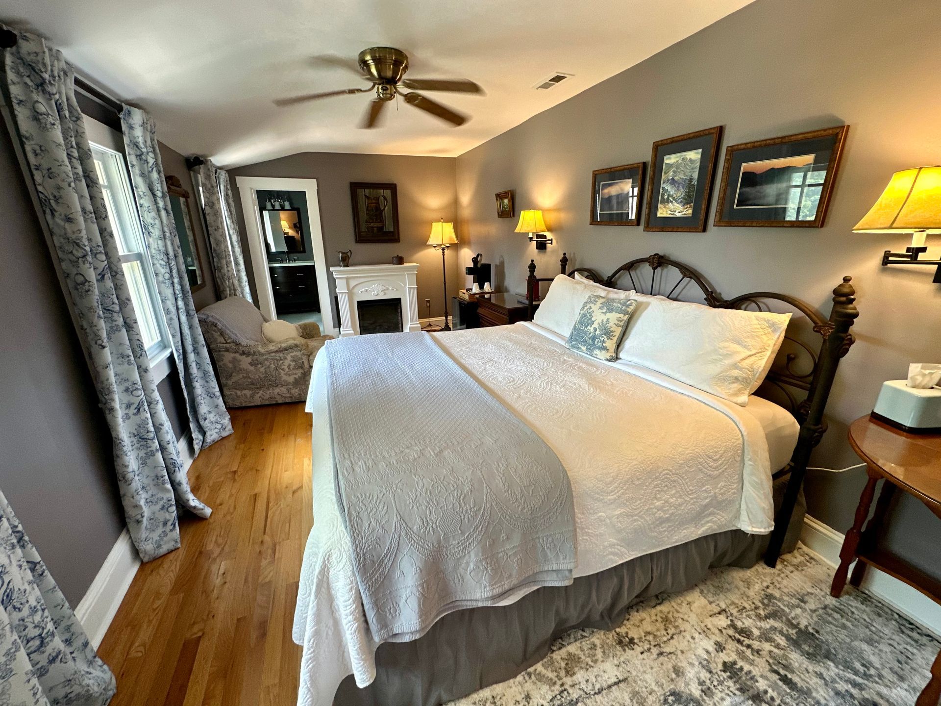 Bedroom with a queen-sized bed, wooden floors, and gray walls. A patterned rug and framed artwork are visible.