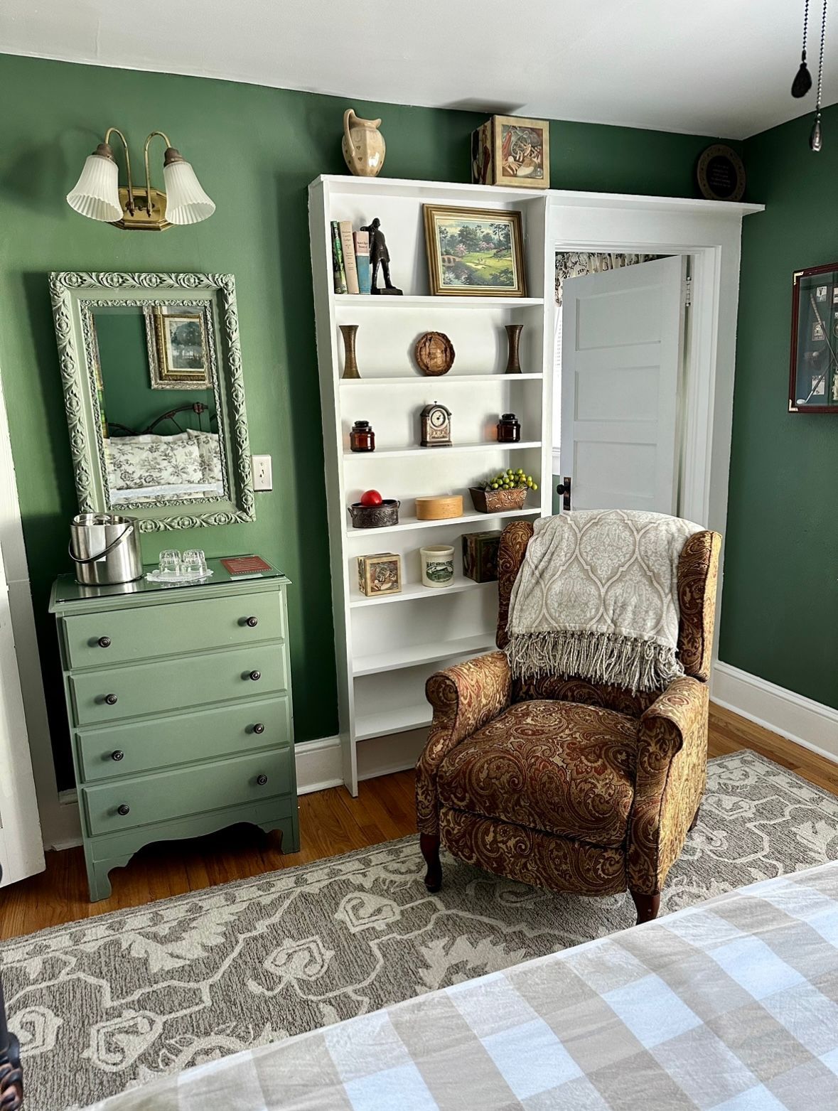 Cozy room with green walls, a patterned armchair, and a white bookcase filled with decorative items. A green dresser and mirror complete the scene.