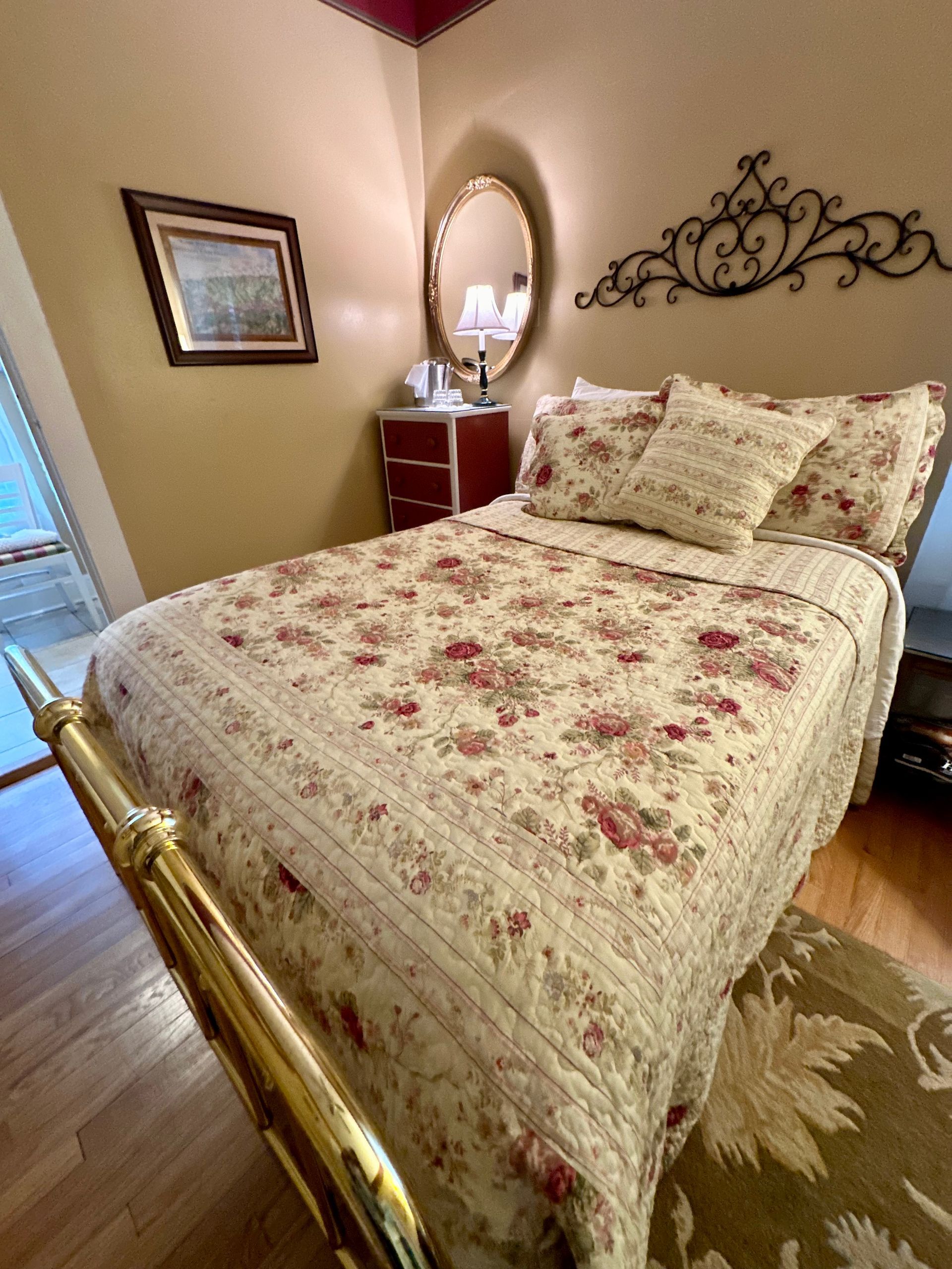 A cozy bedroom with a white lace bedspread and a floral patterned blanket.  A decorative headboard hangs above the bed, with a small dresser and mirror beside it.