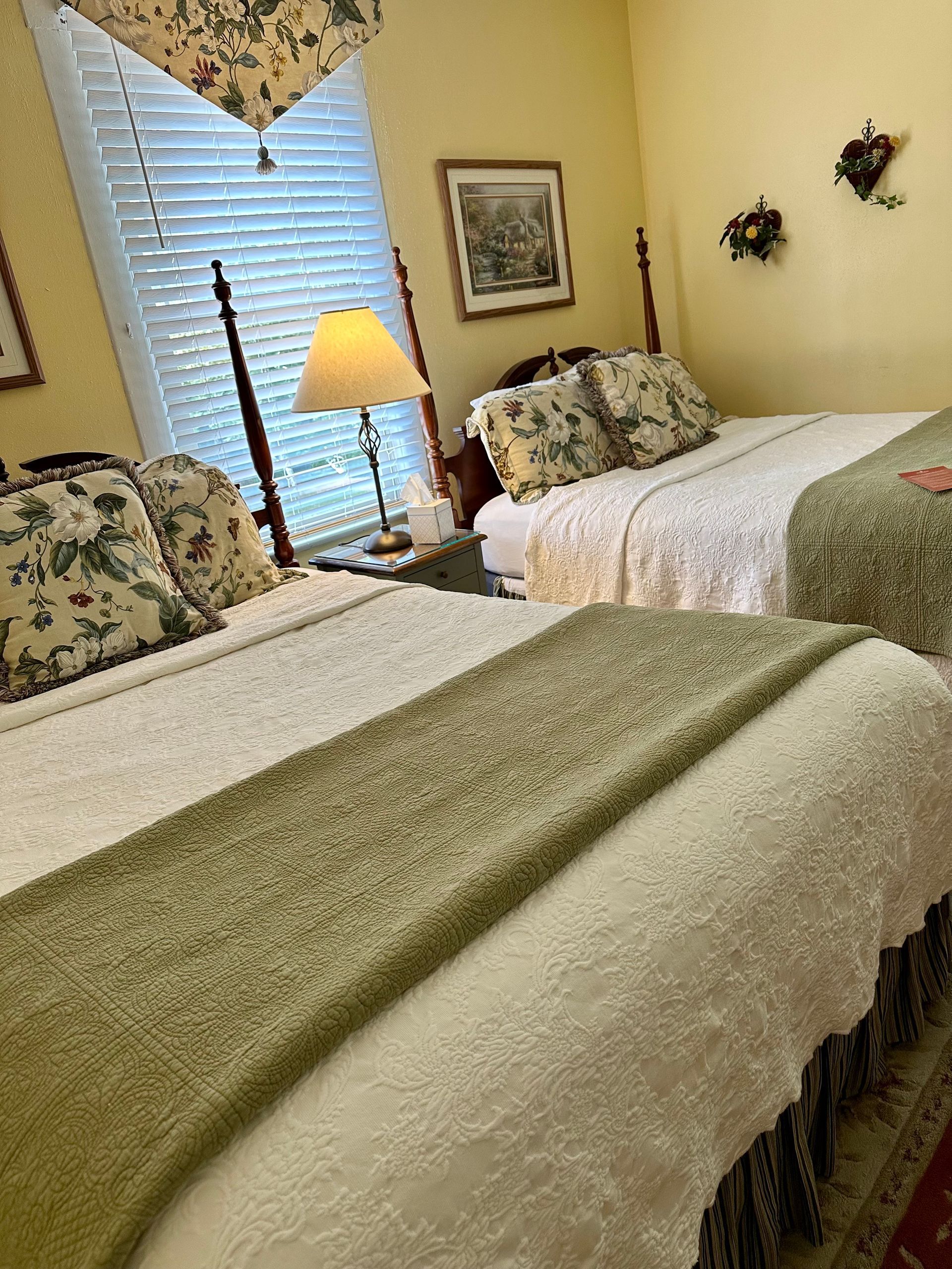 Two beds with floral pillows and white and green bedding in a yellow-walled bedroom. A lamp sits on a nightstand by a window.