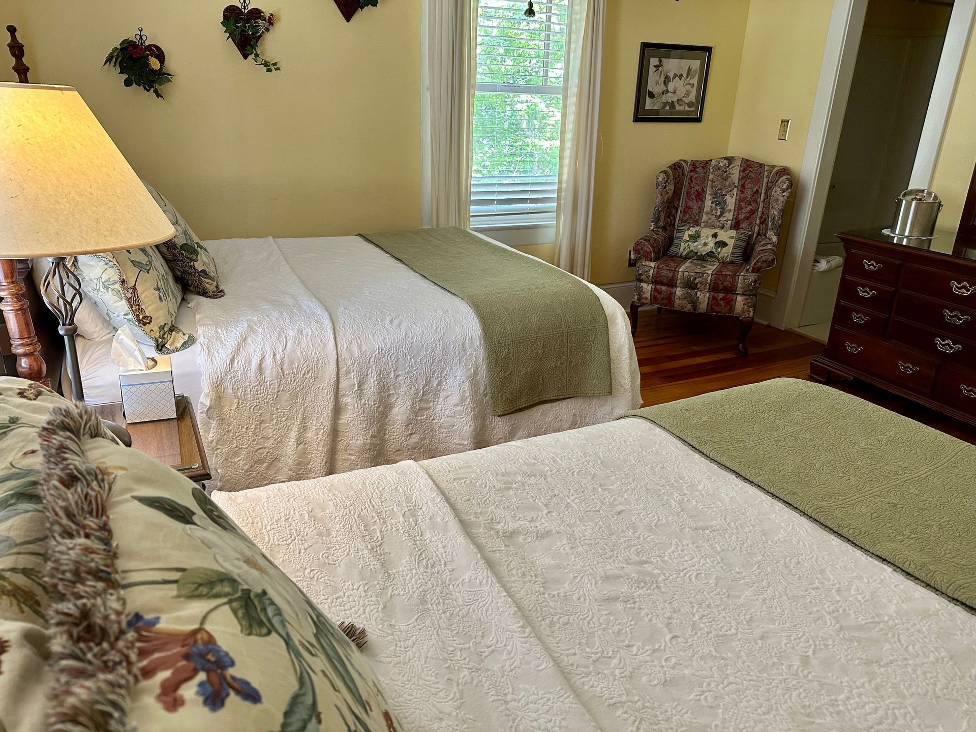 Bedroom with two beds covered in white and green quilts, a floral chair, and a window with a view of trees.