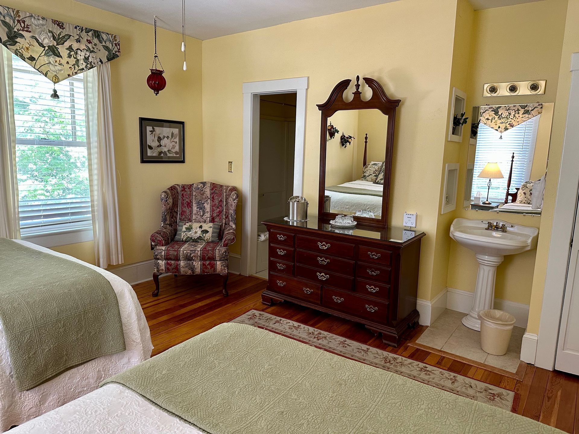 A well-lit bedroom with two beds, a dresser with mirror, armchair, and a sink. The room is yellow with wooden floors.