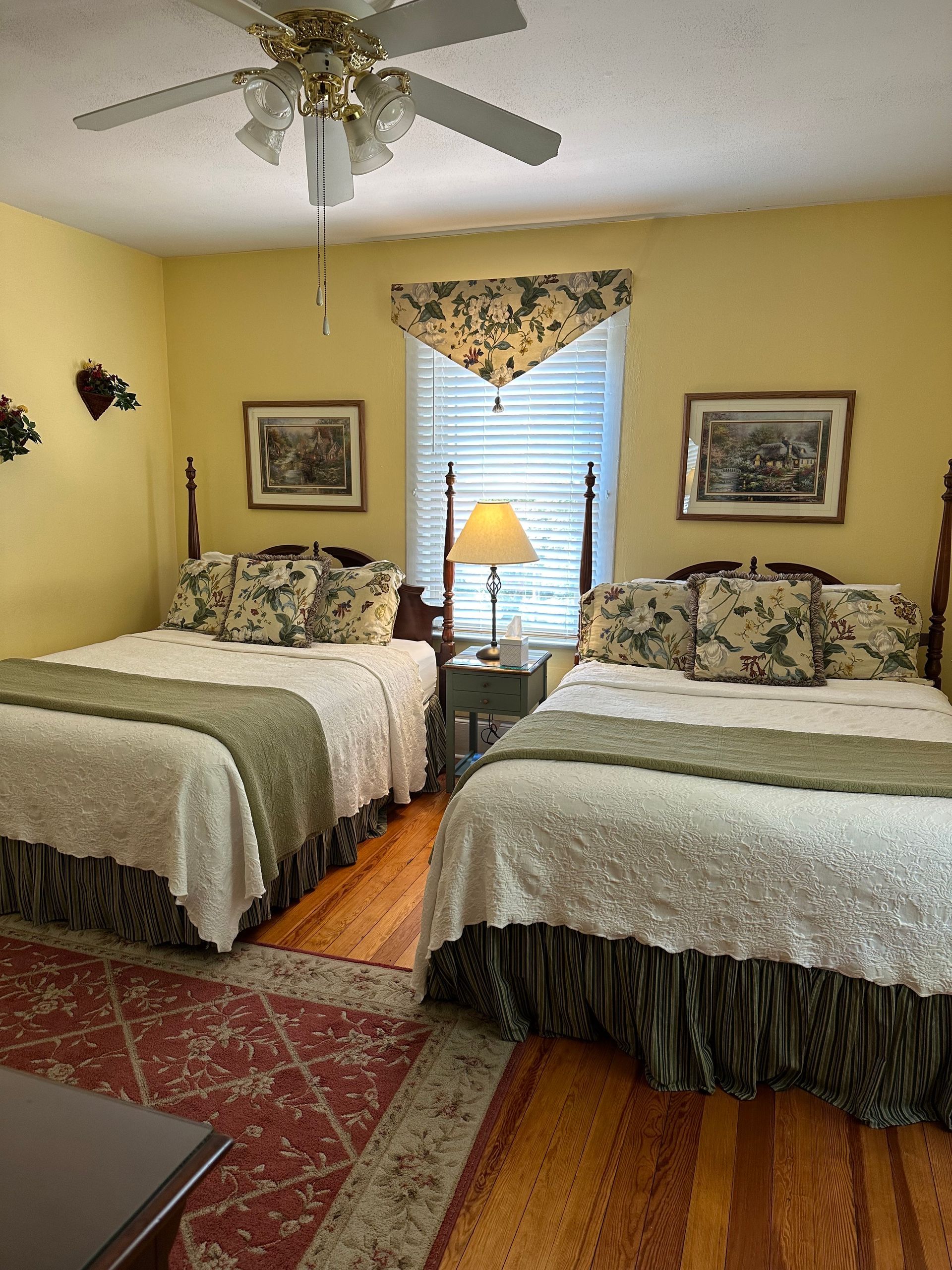 A bedroom with two double beds, yellow walls, wooden floors, and a window with a valance. Each bed has a green and white cover and decorative pillows.