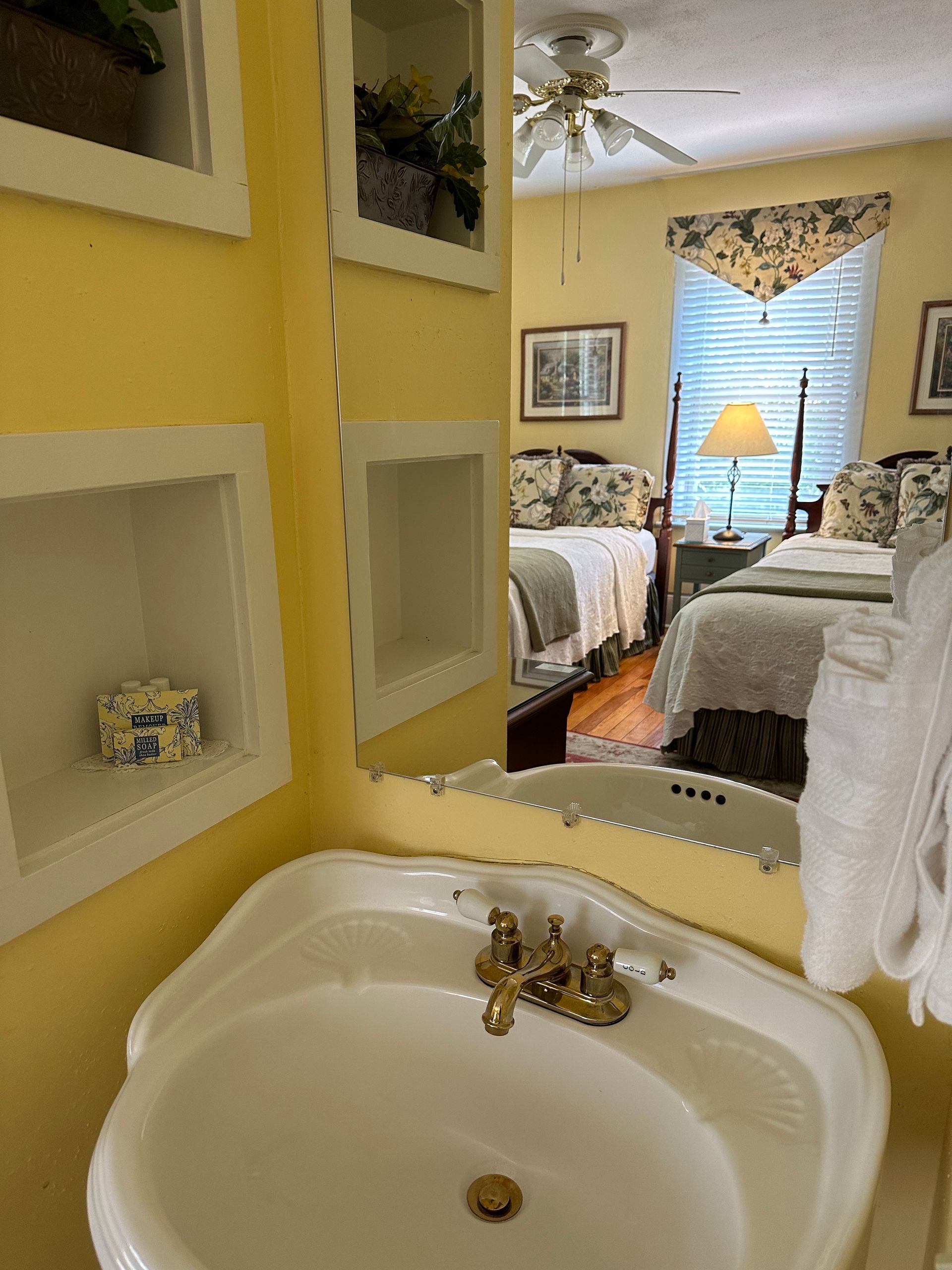 Bathroom with a white sink, gold fixtures, and a mirror reflecting a bedroom with two beds, yellow walls, and built-in shelves.