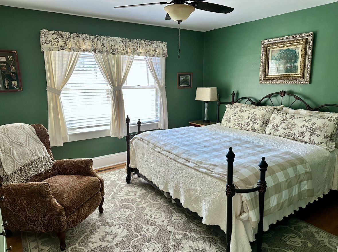Bedroom with a bed, armchair, and window. Green walls, floral bedspread, and ivory curtains.