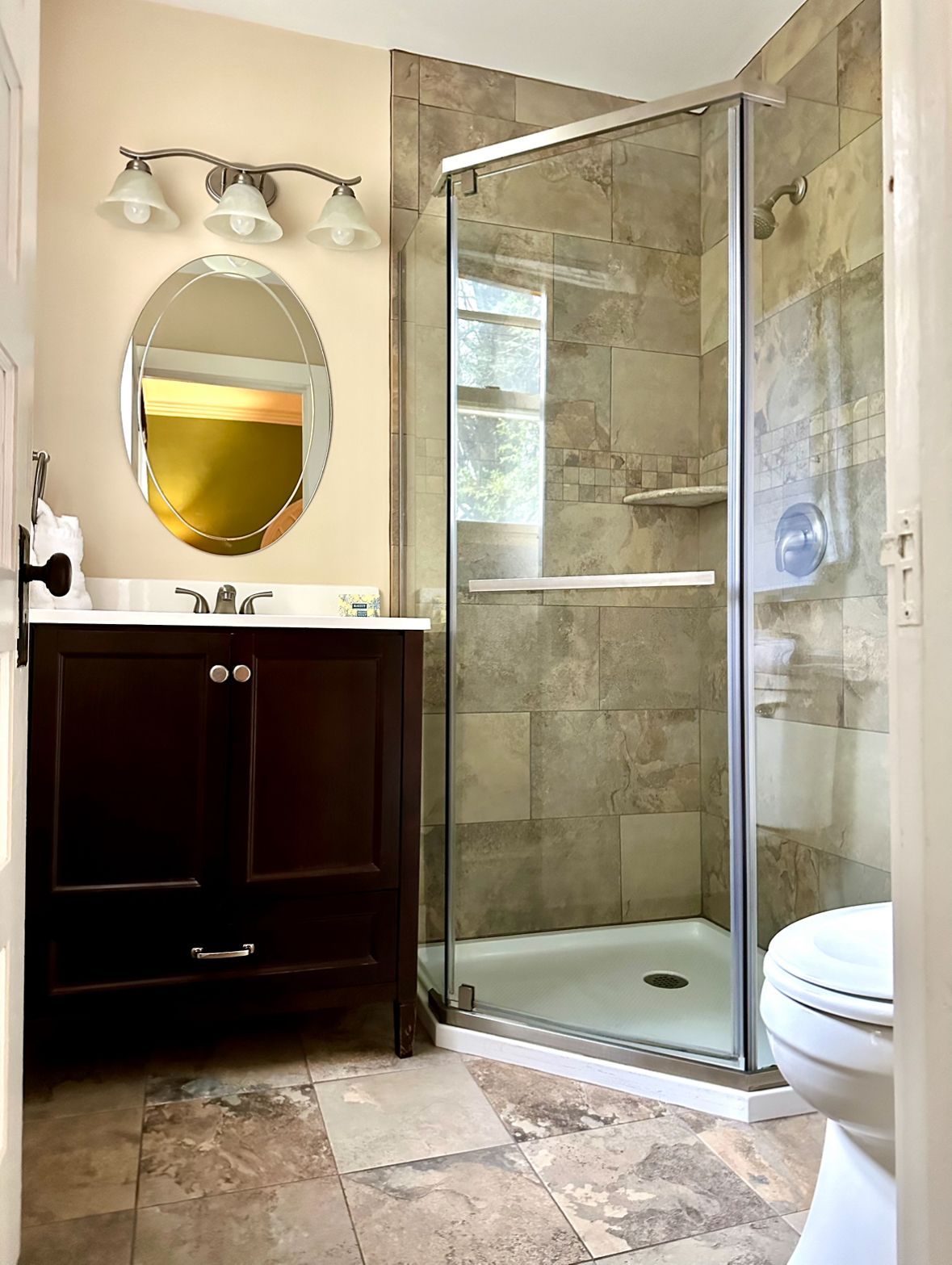 Bathroom with dark brown vanity, oval mirror, glass shower enclosure with beige tiled walls, and a toilet.