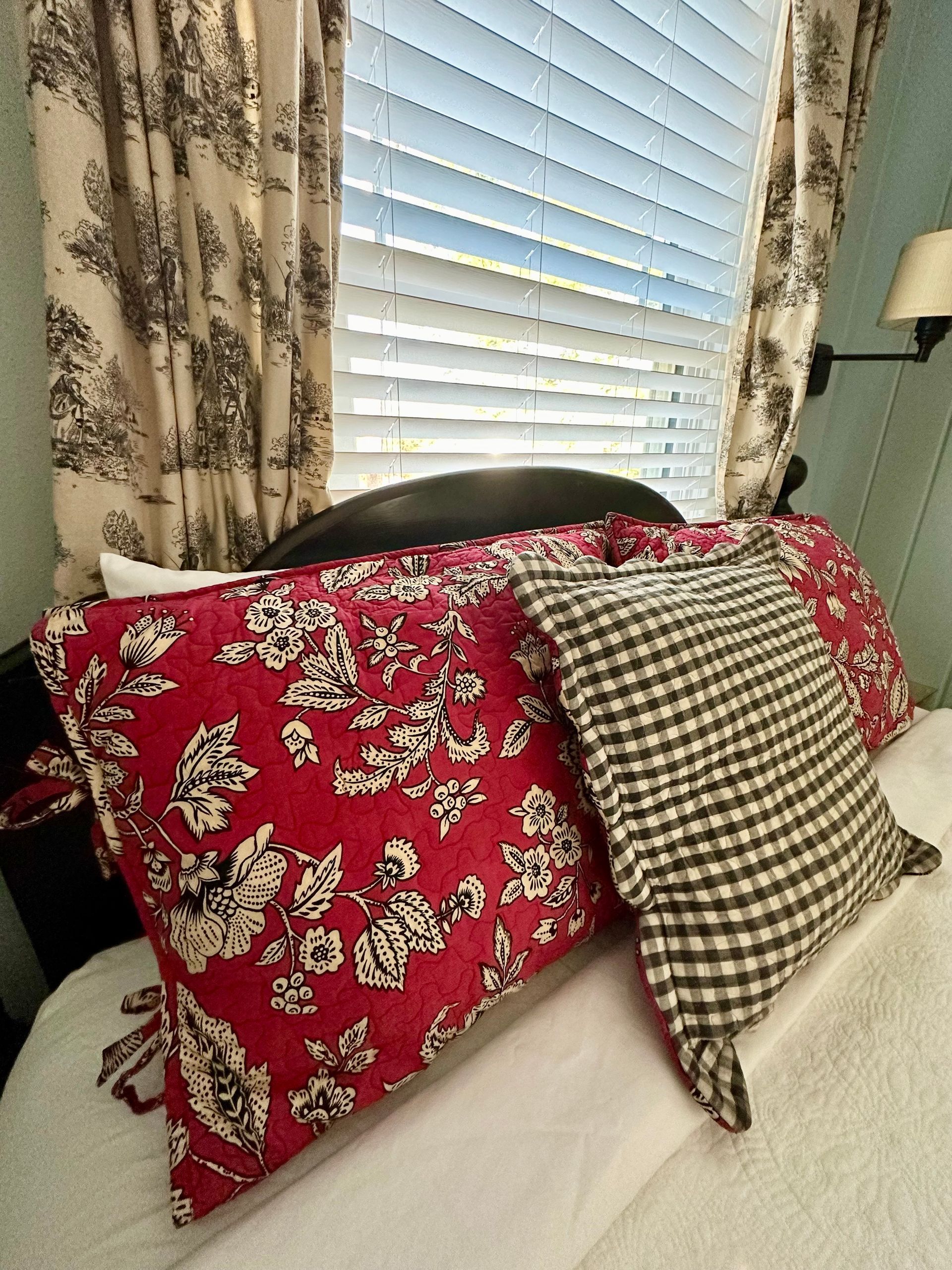 Bed with red floral and black checkered pillows near a window with closed blinds and patterned curtains.
