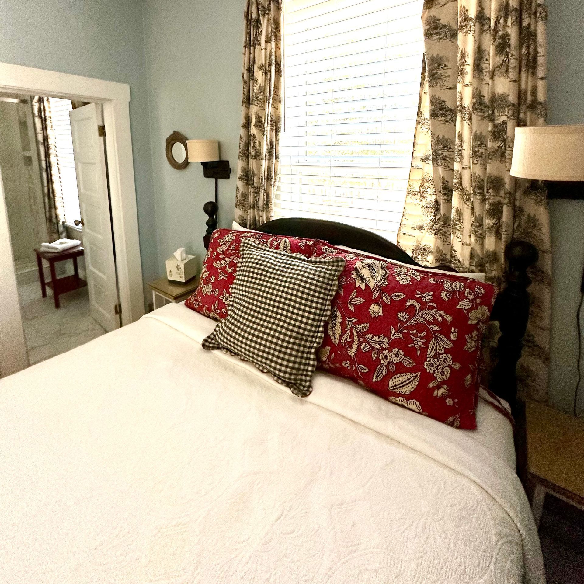 Bedroom with a white bed, red patterned pillows, and a black and white checkered pillow. A window with curtains and a small table are also visible.